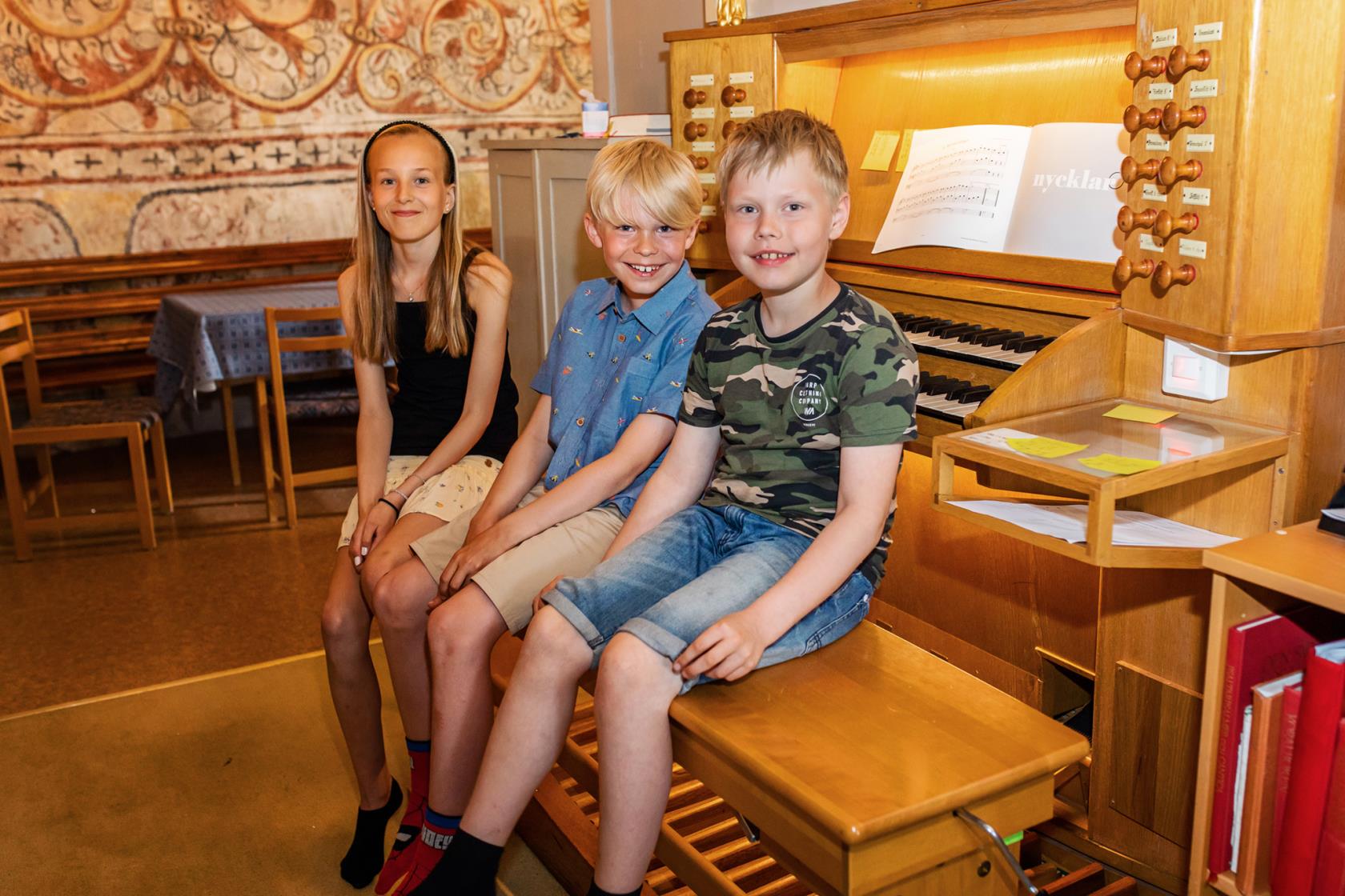 Tre barn, två pojkar och en flicka sitter på en stol framför en kyrkorgel. 