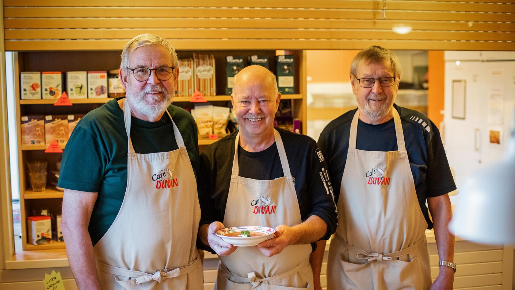 Tre män med förkläden i cafémiljö. Mannen i mitten håller i en tallrik med soppa. 
