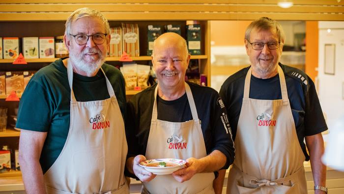 Tre män med förkläden i cafémiljö. Mannen i mitten håller i en tallrik med soppa. 