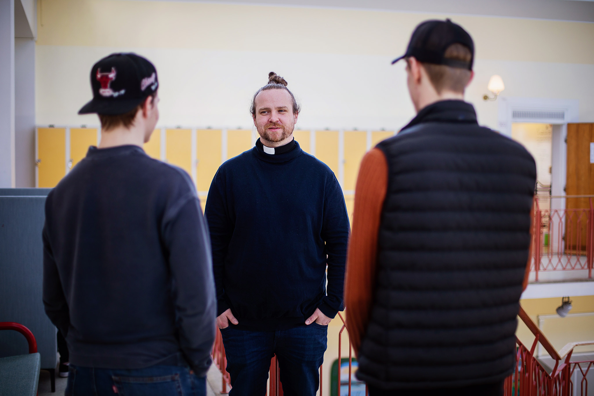 En manlig präst som står i en skolkorridor och pratar med två unga killar som har keps.