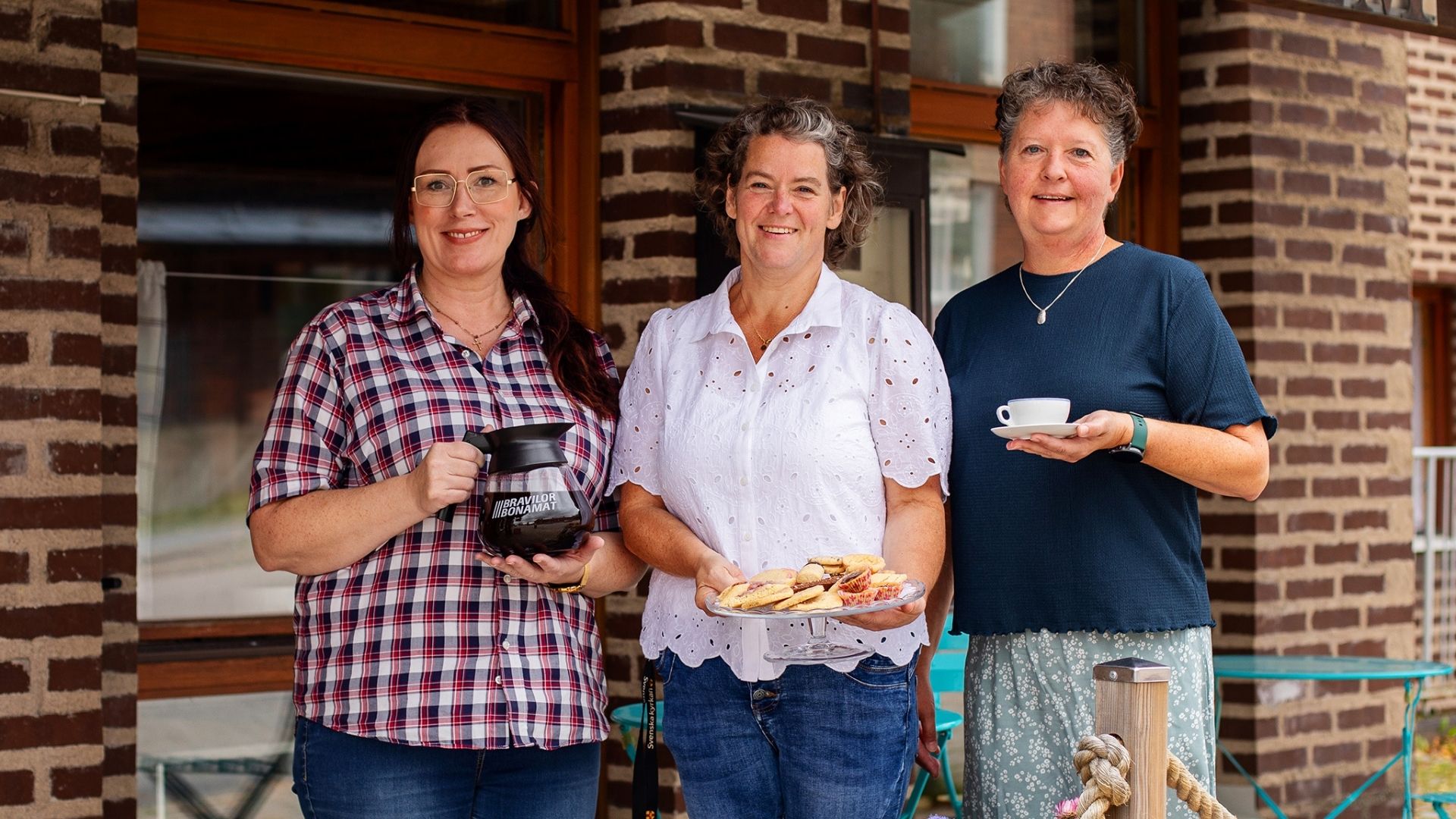 Tre kvinnor som står intill varandra, där en håller i en kaffekopp, en annan en kaffepanna och den tredje håller i ett kakfat.