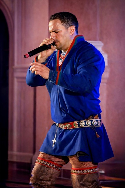 En man klädd i blå samisk kolt, bälte runt midjan och skinnbyxor. Han håller en mikrofon i sin hand och sjunger. 