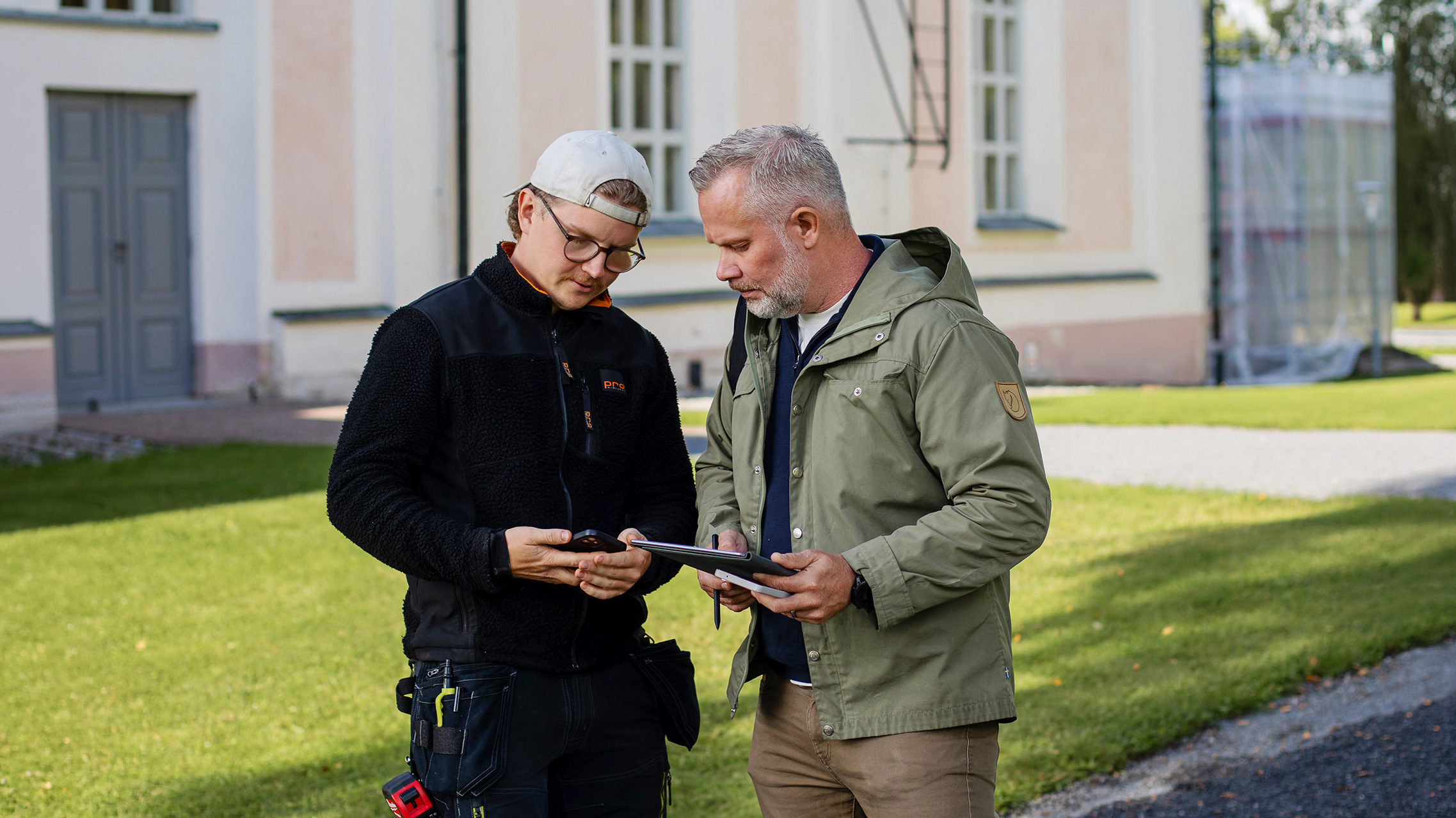 Två män som står och pratar med varandra framför en rosa byggnad. Den ena mannen håller i en läsplatta och den andra en mobiltelefon.