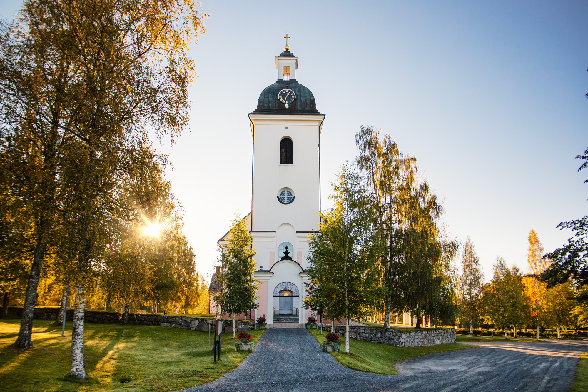 En frasida på en kyrkbyggnad, där man ser kyrkporten. I bakgrunden skiner solen genom träden.