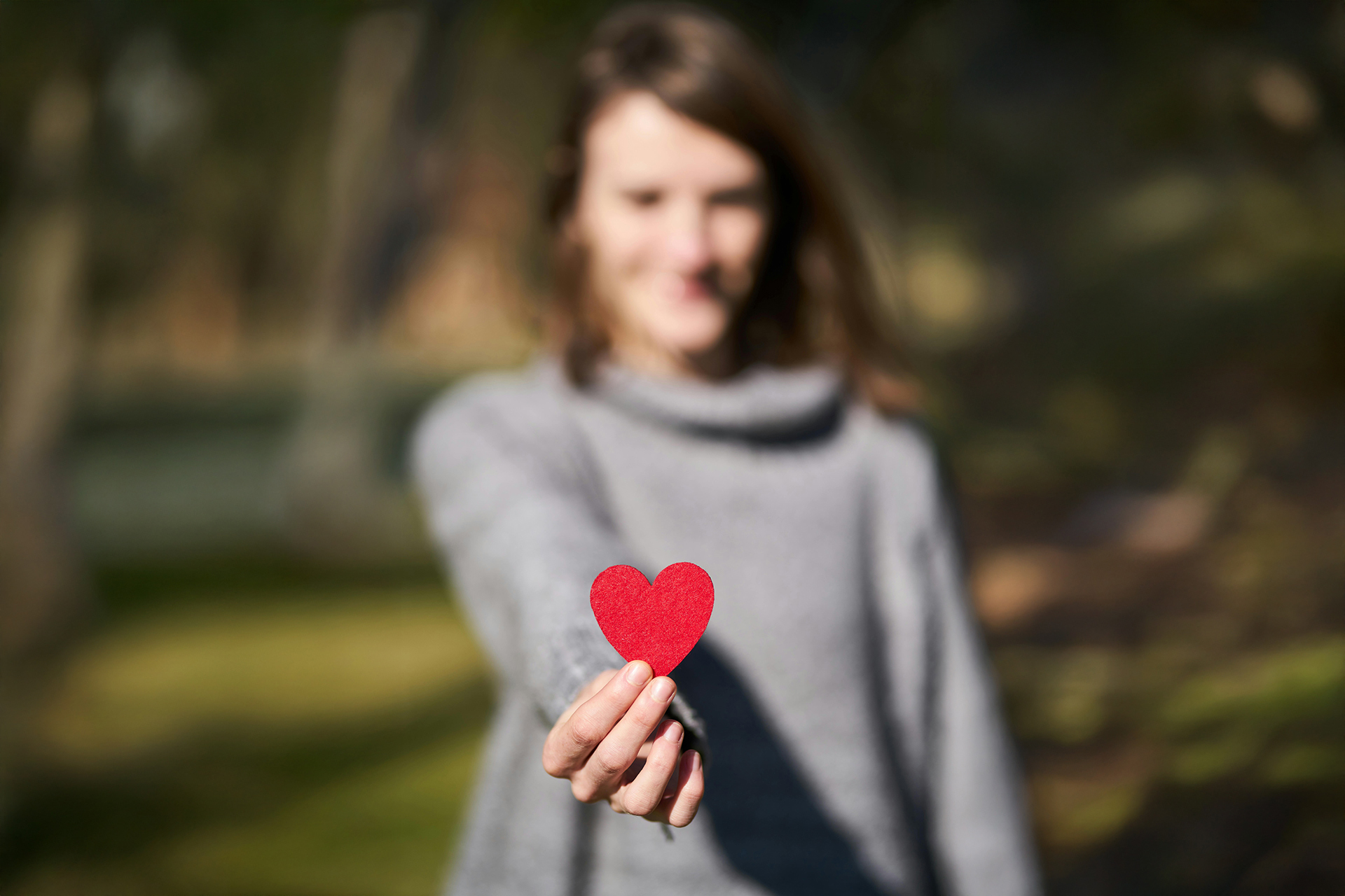 Kvinna med brunt hår och grå tröja sträcker ut en hand där hon håller ett litet rött hjärta.