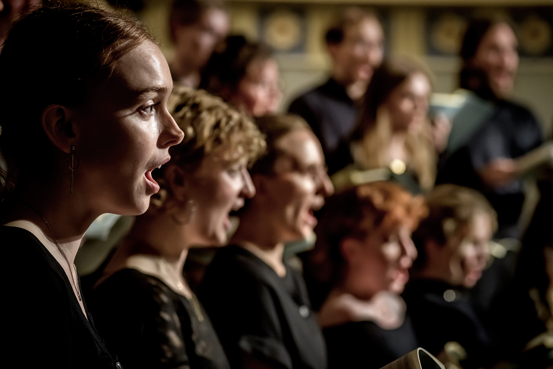 Ett antal körsångare i mörka kläder syns i profil sedda från höger i full färds med att sjunga ut.