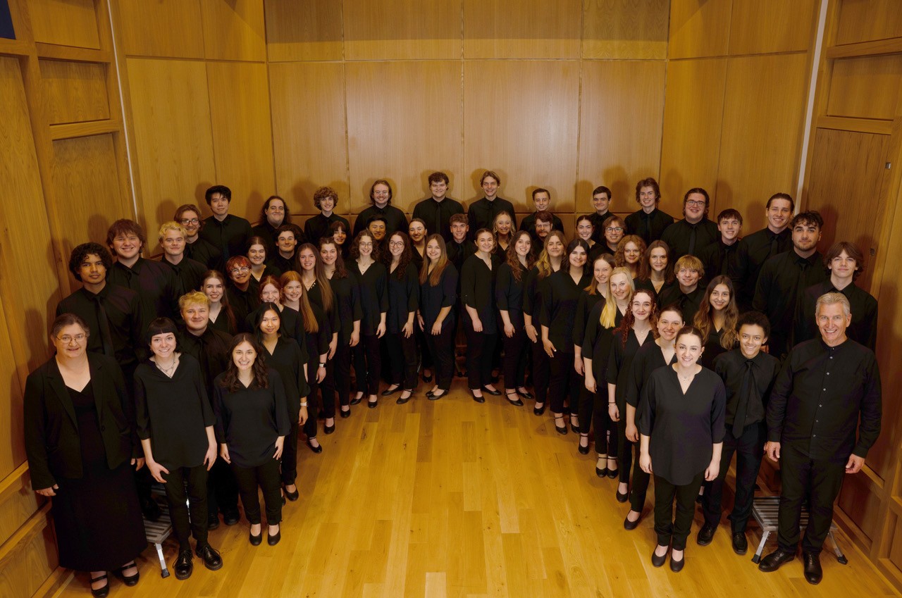 Augustana Choir står uppställda i en tre raders halvcirkel, klädda i svart, i ett rum med trägolv och träväggar.