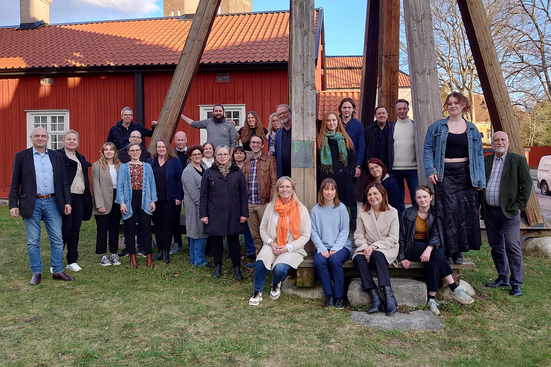 Kören Camerata Incantus står uppställd vid en klockstapel i närheten av Västerås domkyrka med blönaddad klädsel och en faluröd byggnad i bakgrunden.