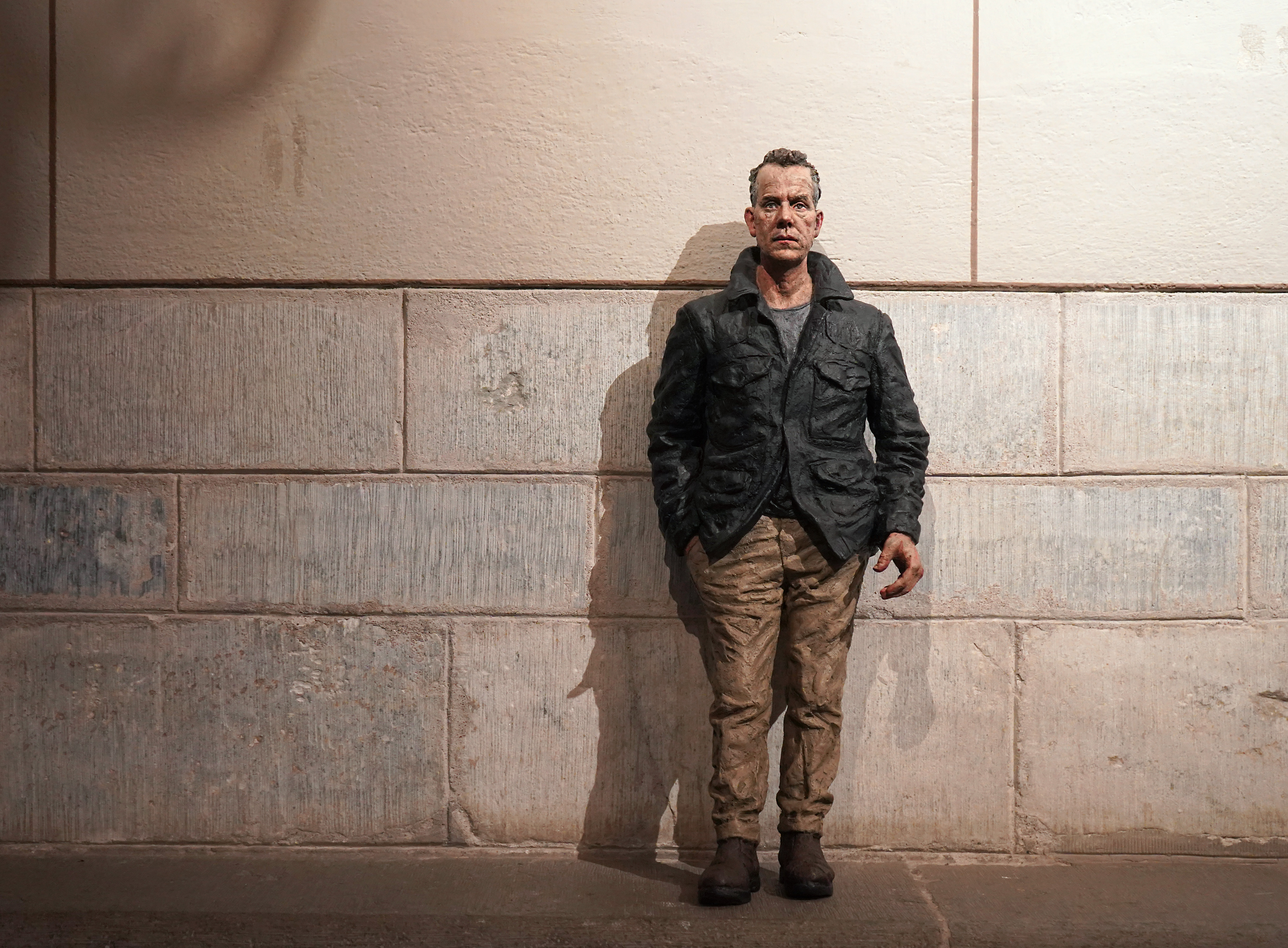 En manslik skulptur i brons med skrynkliga kläder i jordiga nyanser står lutad mot väggen i Uppsala domkyrkas södra koromgång och spänner blicken i tittaren med ena handen i byxfickan.