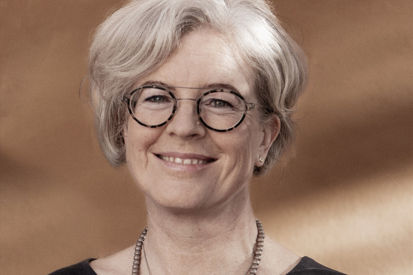 Ewa Krtmeijer, med glasögon, halsband och grå, kortklippt frisyr, på klassisk porträttbild mot lattebrun bakgrund.