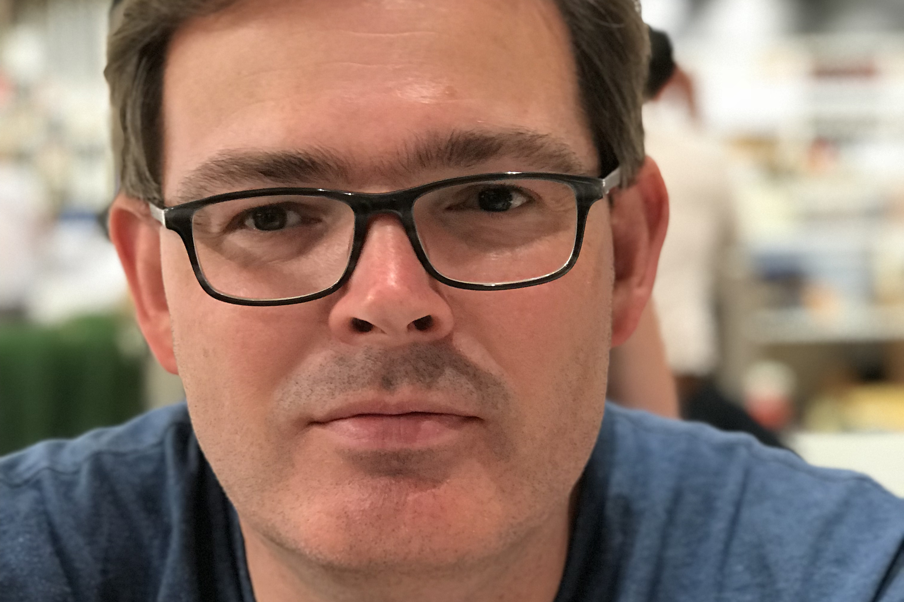 Anthony Gritten, i blå tröja och svartbågade glasögon, möter betraktarens blick via närgången porträttbild.