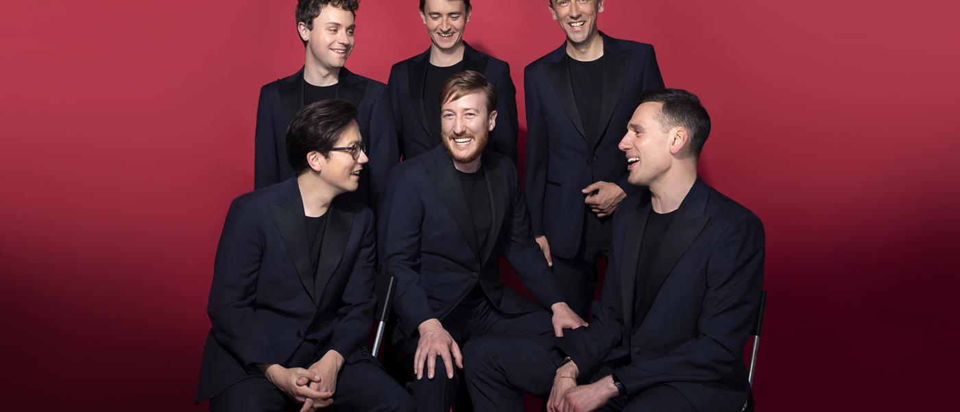 Sångsextetten King's Singers, sex män i mörk kostym mot röd bakgrund, spexar och skrattar på ett klassiskt gruppfoto.
