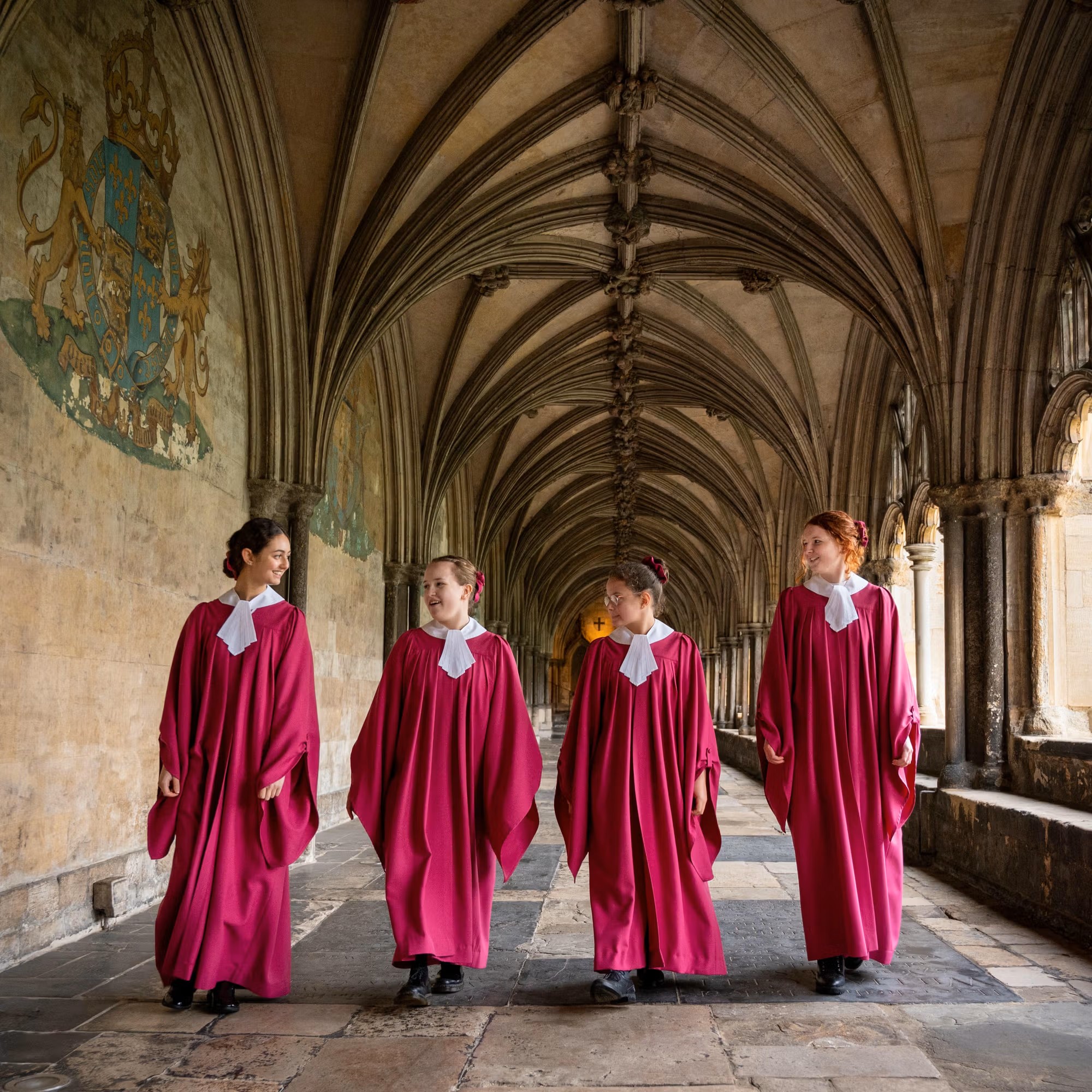 Fyra kvinnliga korister i ceriseröda kåpor går tillsammans under valven i vad som ser ut att vara klassisk engelsk kyrkomiljö i Norwich Cathedral.