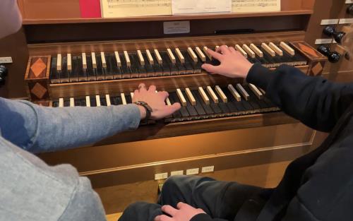 En organist och en orgelelev sitter ner tillsammans vid Helga Trefaldighets kyrkas orgel och spelar tillsammans på manualernas tangenter.