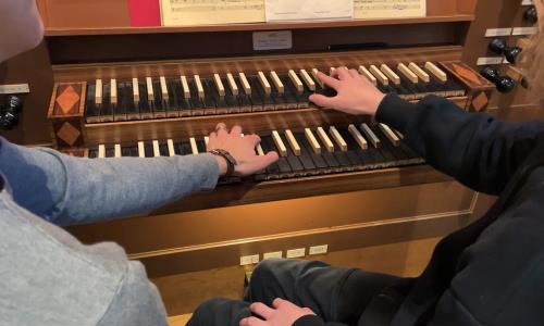 En organist och en orgelelev sitter ner tillsammans vid Helga Trefaldighets kyrkas orgel och spelar tillsammans på manualernas tangenter.