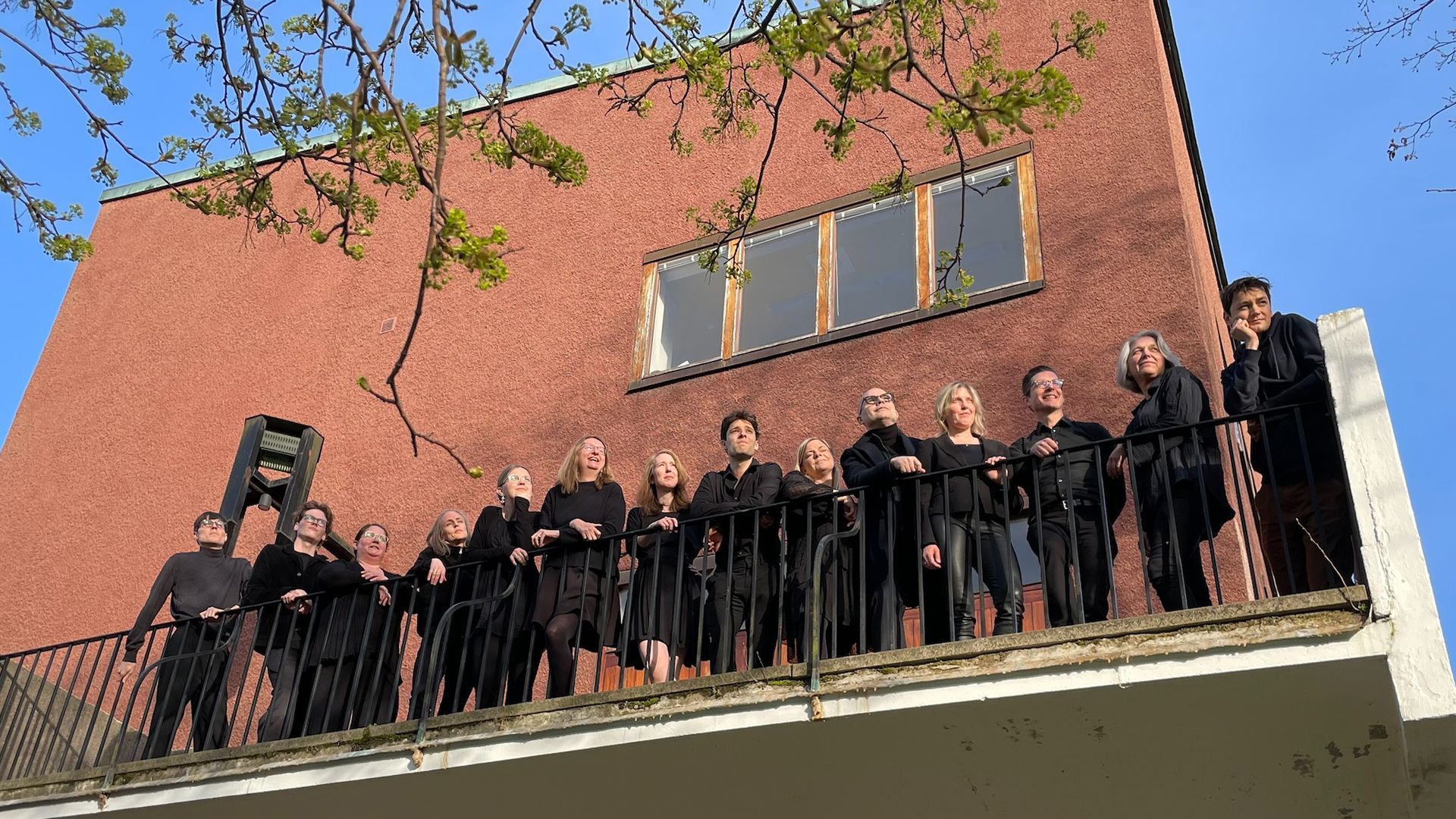 Oscarskyrkans vokalensemble står svartklädda och uppradade längs ett balkongräcke med blå himmel i bakgrunden och blickarna ner mot betraktaren/fotografen.