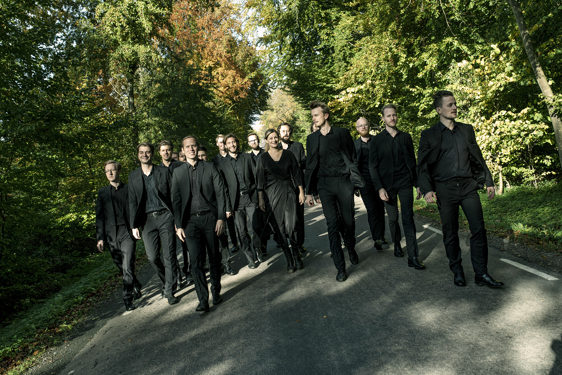 Kören Svanholm singers promenerar vägen fram i en höstfärgad skogsglänta, iförda svarta kostymer, på väg mot betraktaren.