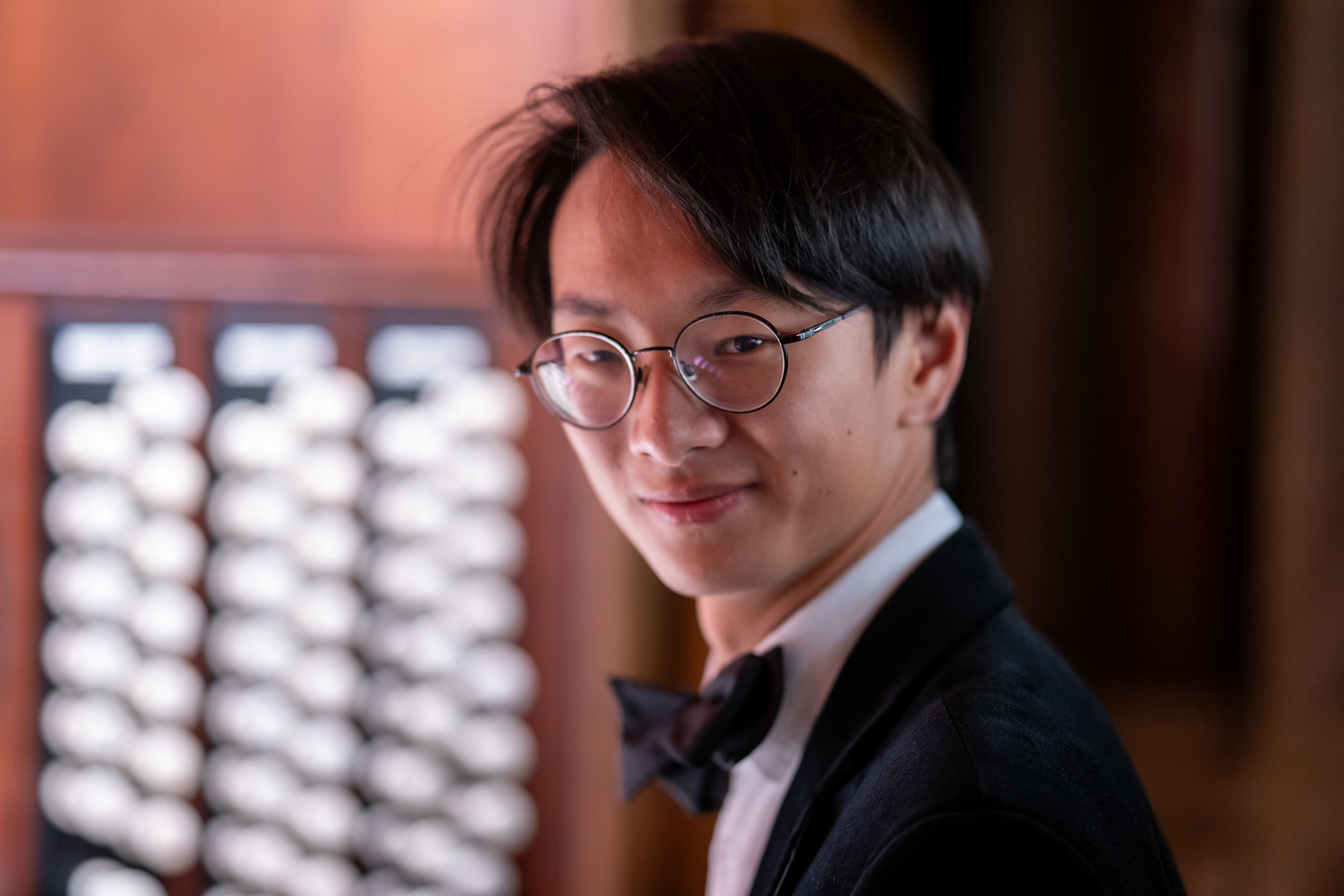 Tingshuo Yang i kostym och svart fluga samt runda glasögon ler undefundigt under lugg mot betraktaren. I bakgrunden syns registerpanelen på en kyrkorgel.