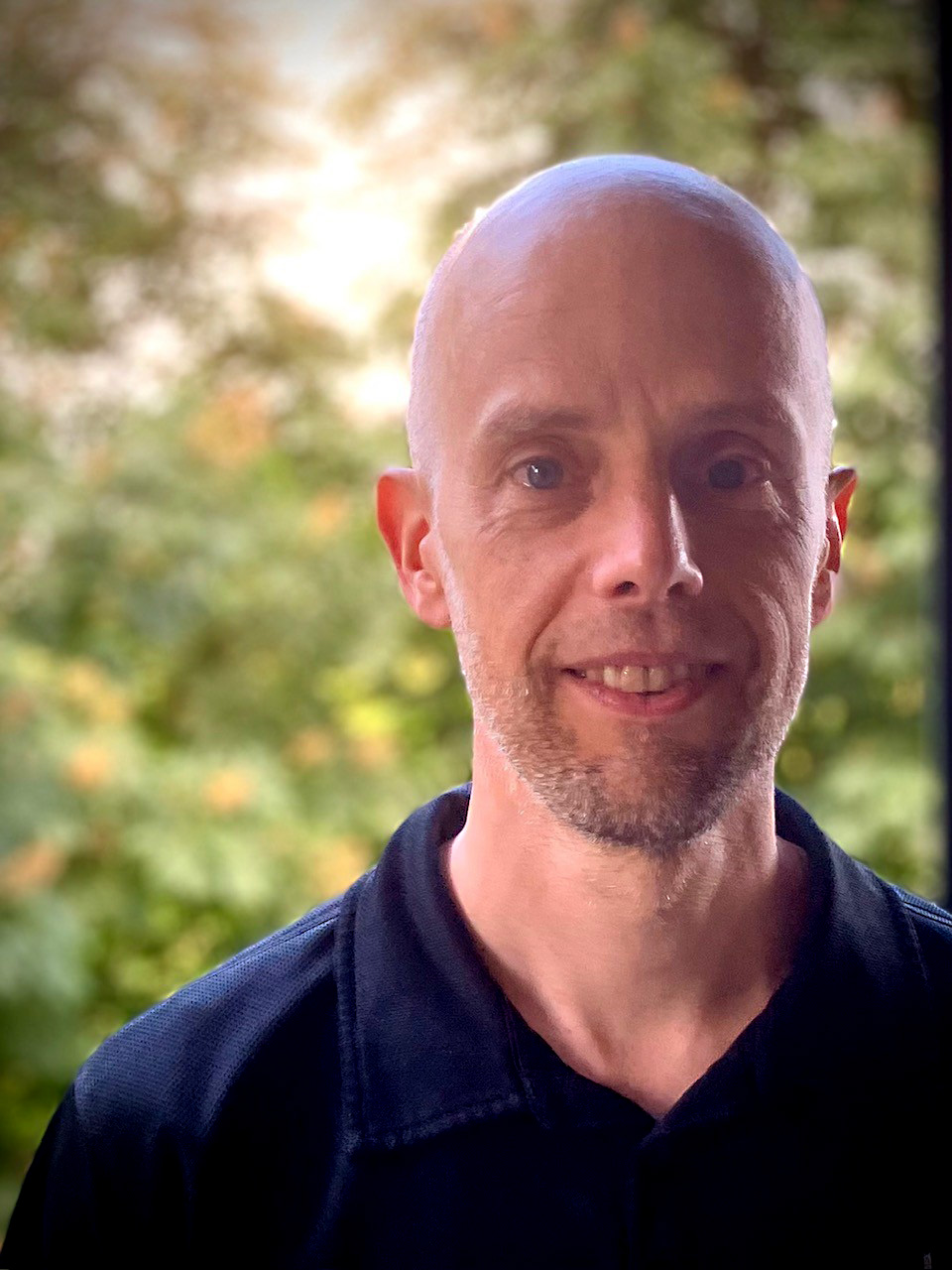 Tomas Jönson i blå skjorta, avporträtterad i motljus mot grönskande och solig bakgrund.