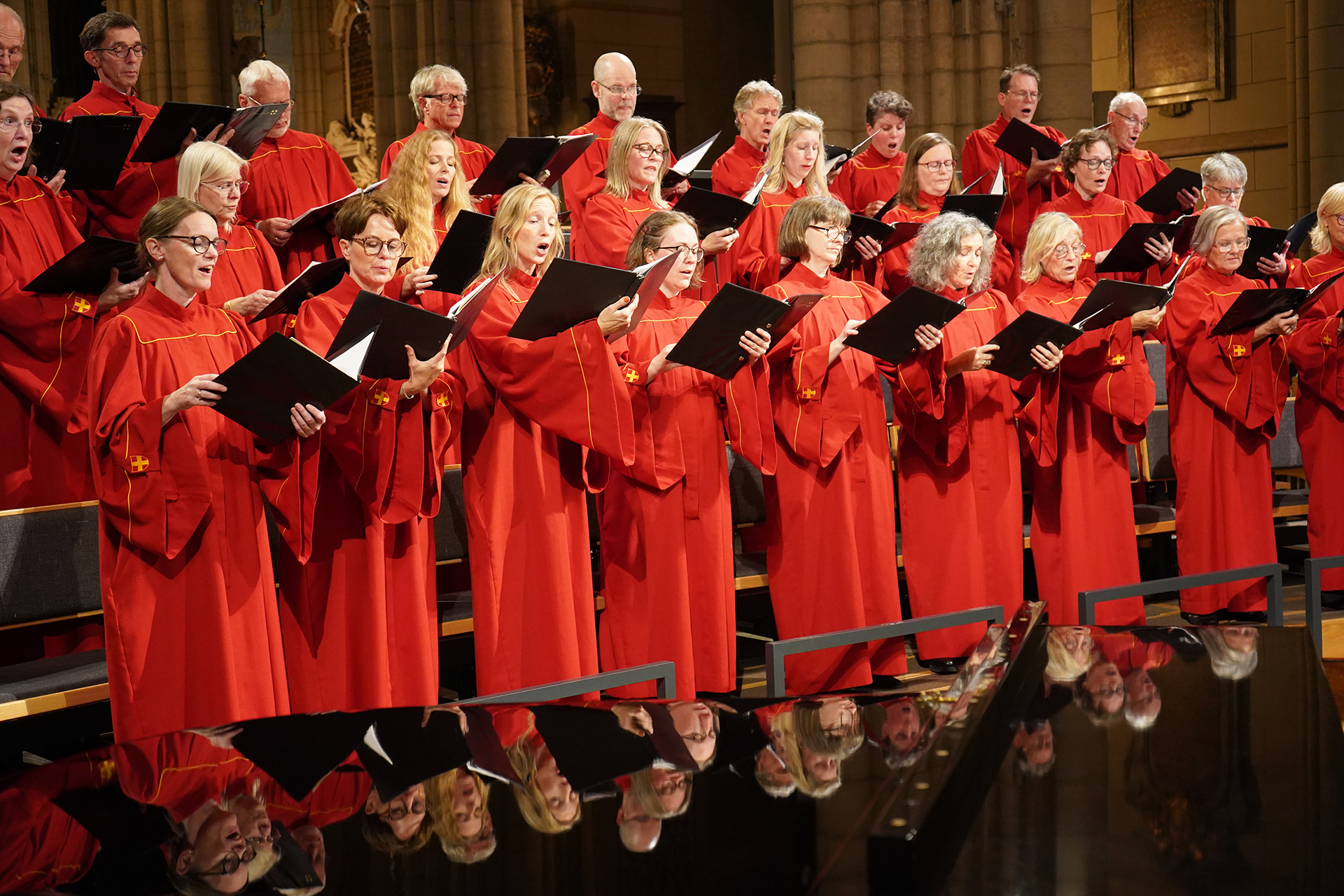 Uppsala domkyrkokör i röda kåpor sjunger och avspeglas i ovansidan på en blank flygel.