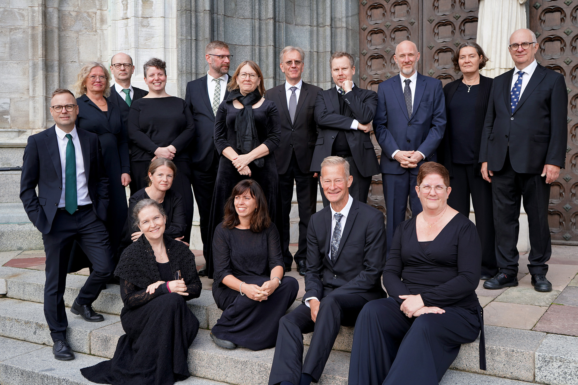 Uppsala katedralsångares korister står och sitter på domkyrkans södra trappa i mörka kläder och avslappnat utspridda.