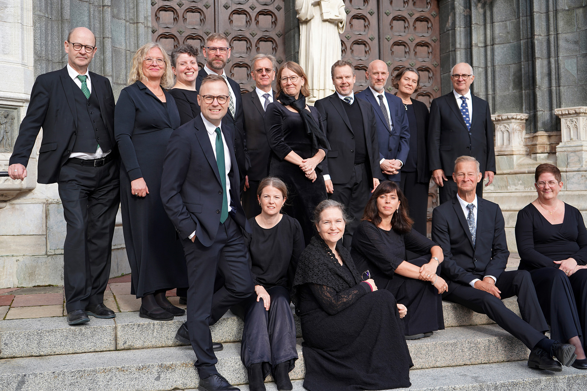 Uppsala katedralsångare, klädda i svart, avslappnat grupperade sittande och stående på trappan vid Uppsala domkyrkas södra port..
