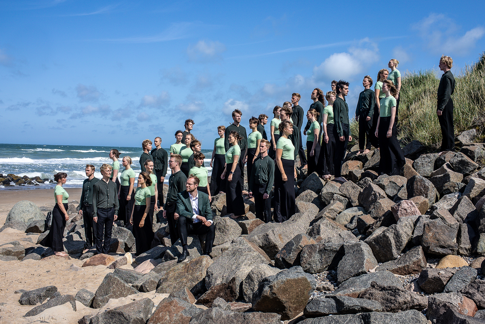 Ungdomskören Aarhus U står uppställda på en strand med blå himmel och vågor med vita gäss i bakgrunden, huller om buller i ett parti med stenar. Kvinnorna är klädda i svarta byxor och mintgröna tröjor, männen i mörka byxor och mörkgröna skjortor.