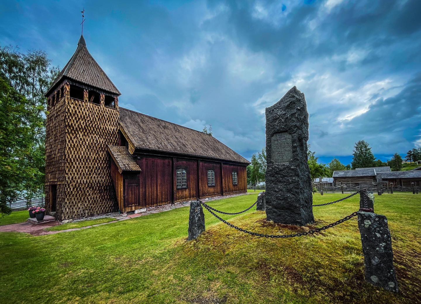 Gemmal träkyrka med grönt gräs runt om och en minnessten i förgrunden