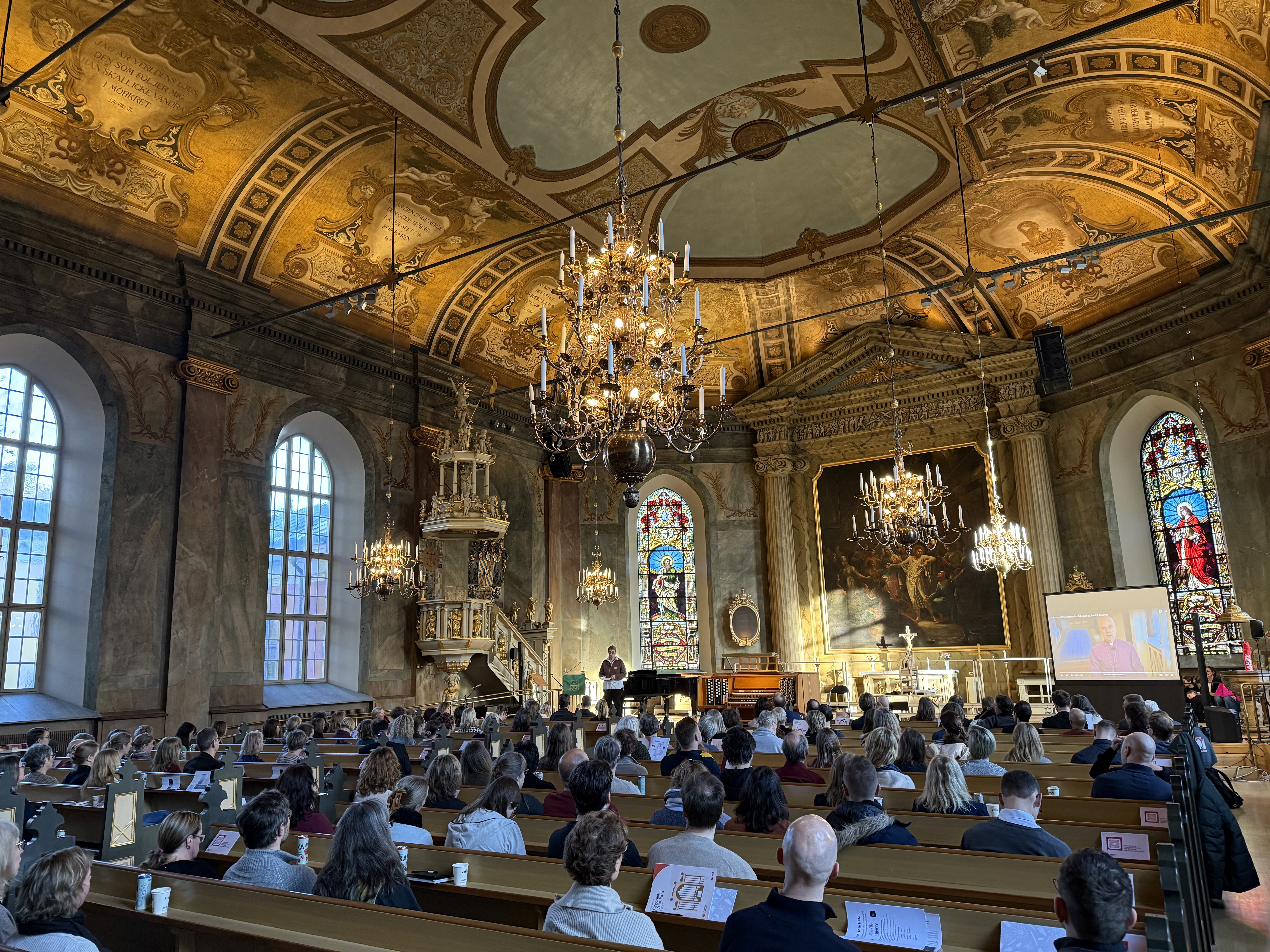 En fullsatt kyrkosal där takmålningarna lyser gult.