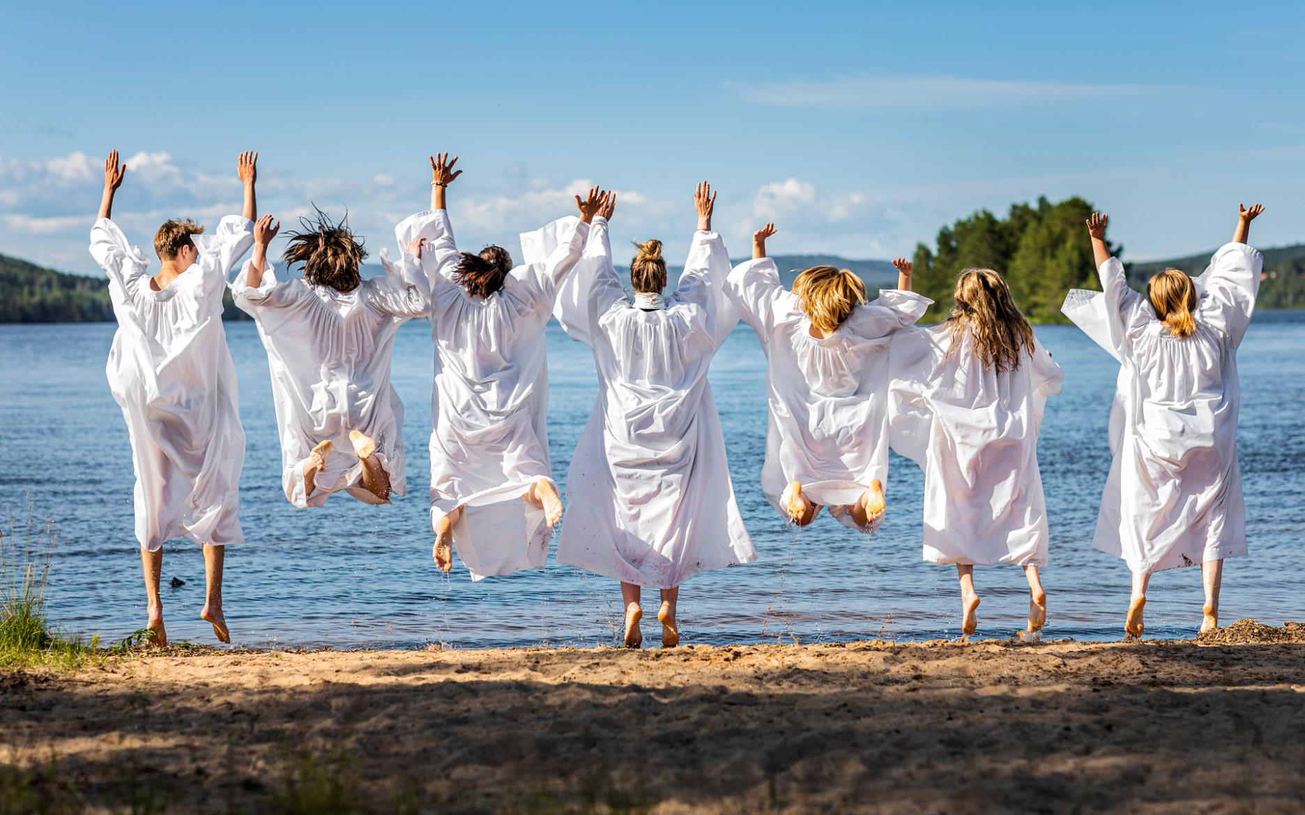 Sju ungdomar i vita konfirmationskåpor hoppar upp i luften. De befinner sig på en sandstrand med havet i bakgrunden.
