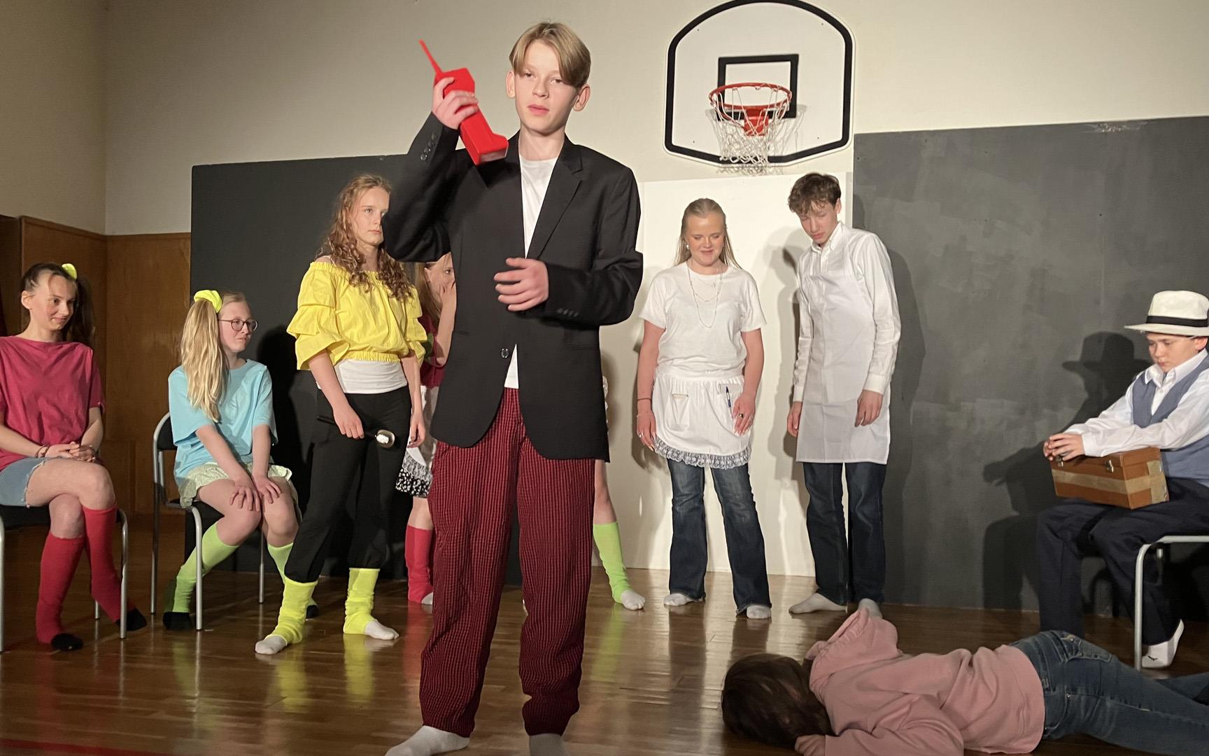 Tio ungdomar som spelar teater. En person ligger på golvet, i förgrunden står en person med en gammaldags röd telefon i handen. Övriga i bilden tittar på personen som ligger på golvet.