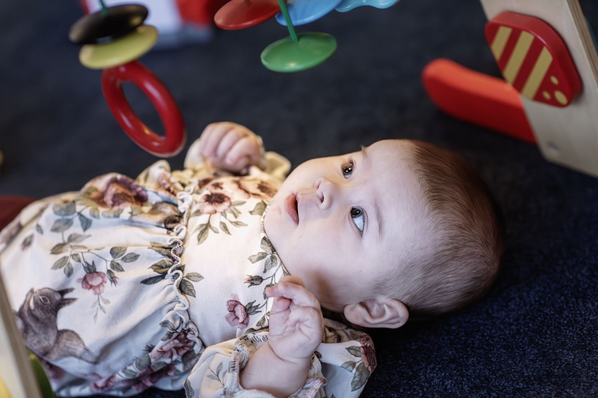 En liten tjej ligger på golvet och tittar upp mot en mobil med hängande leksaker.