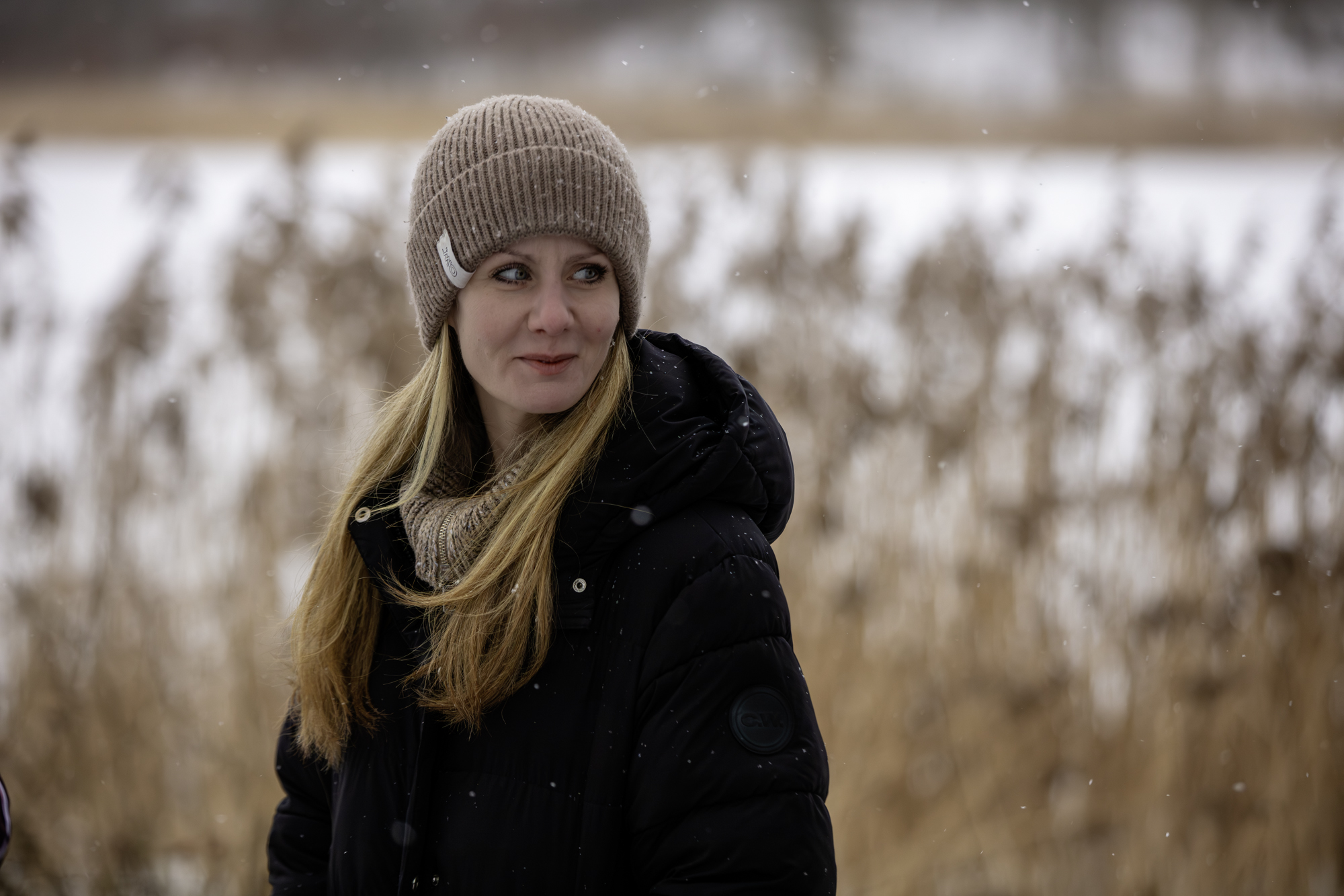 Tjej med långt blont hår och vinterjacka och mössa. Vass och sjö i bakgrunden. per