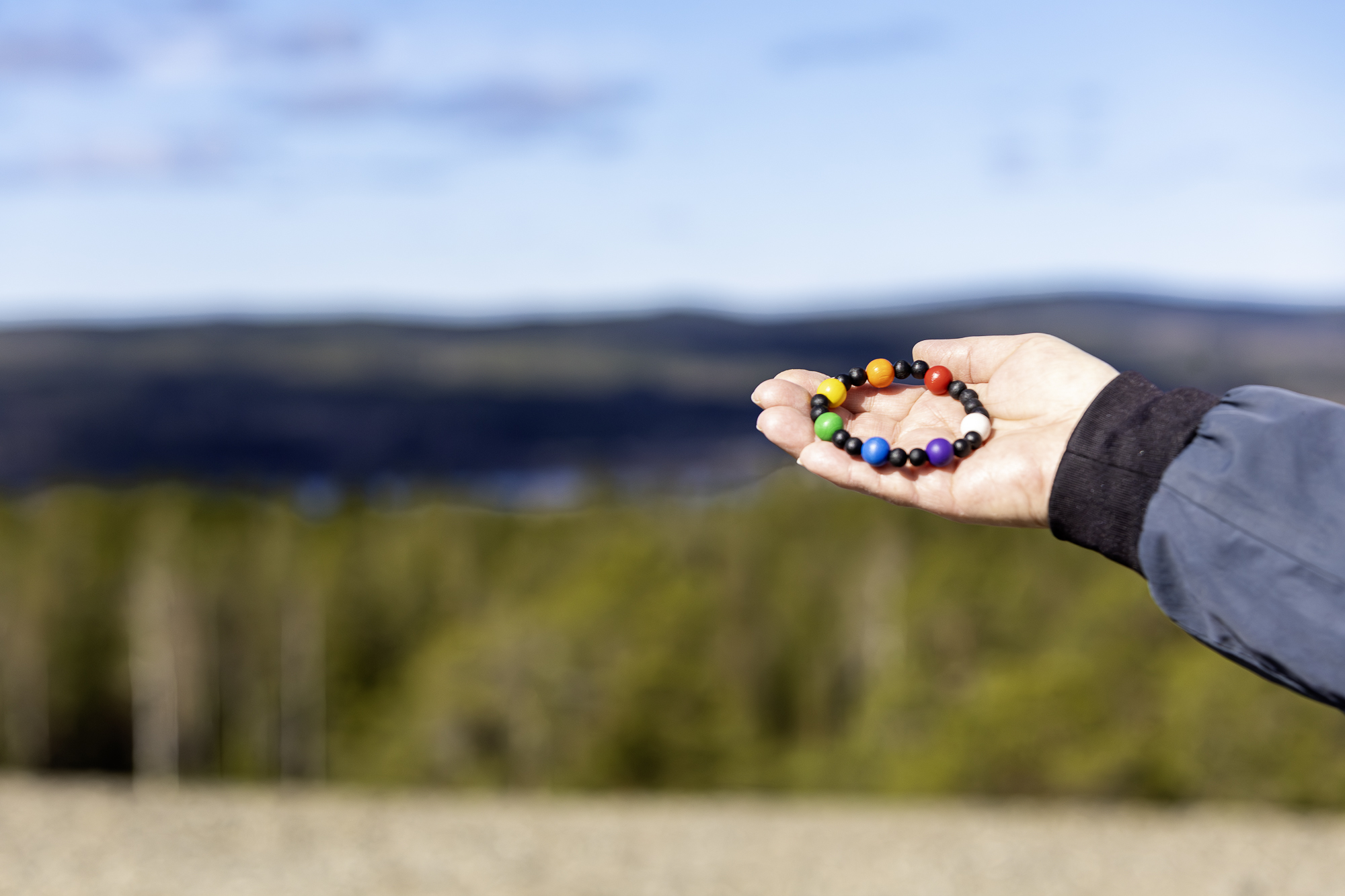 En hand håller upp ett pilgrimsarmband, vacker utsikt över skog och vatten