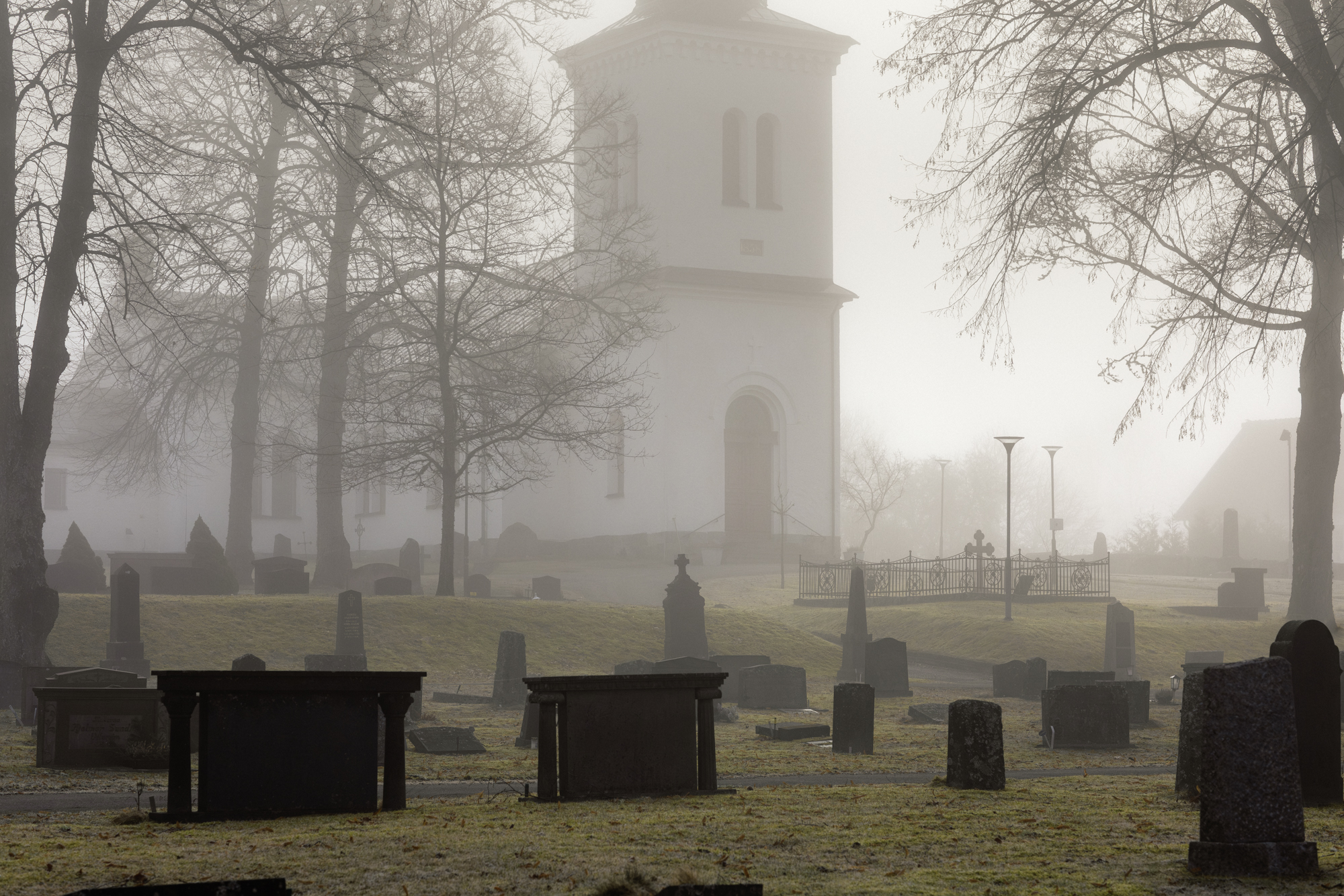 Näsby kyrkogård en dimmig morgon. Mörka gravstenar syns i rader framför kyrkan. 
