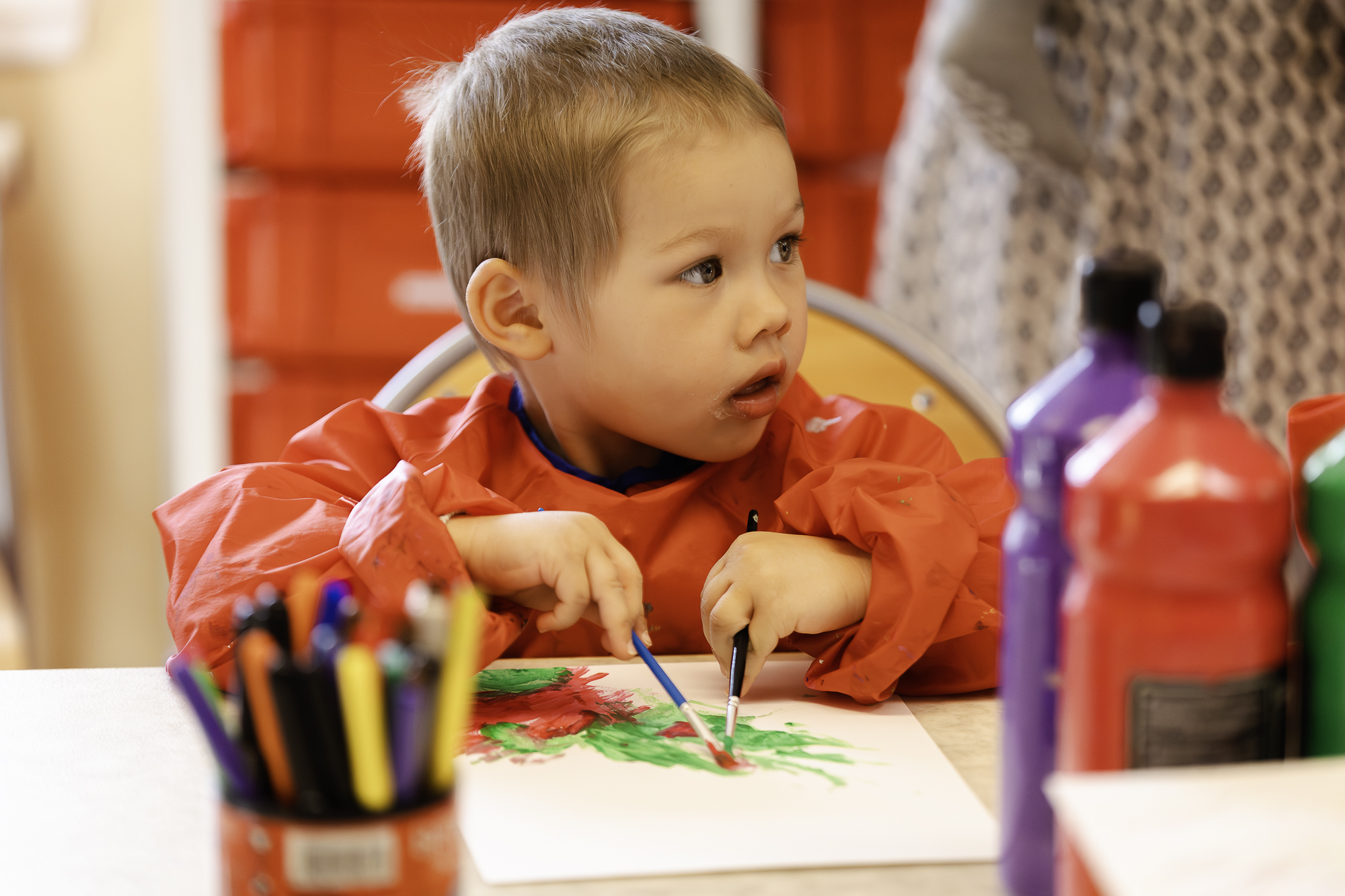 En pojke sitter med målarförkläde och en pensel i varje hand och målar