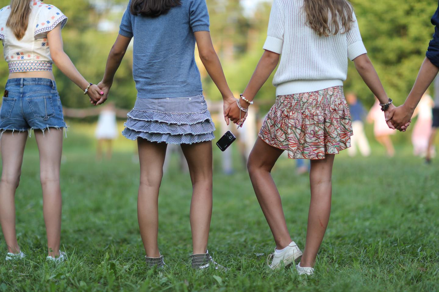 Tonårstjejer i kjol, ståendes i grönt somargräs, håller varandra i händerna. 