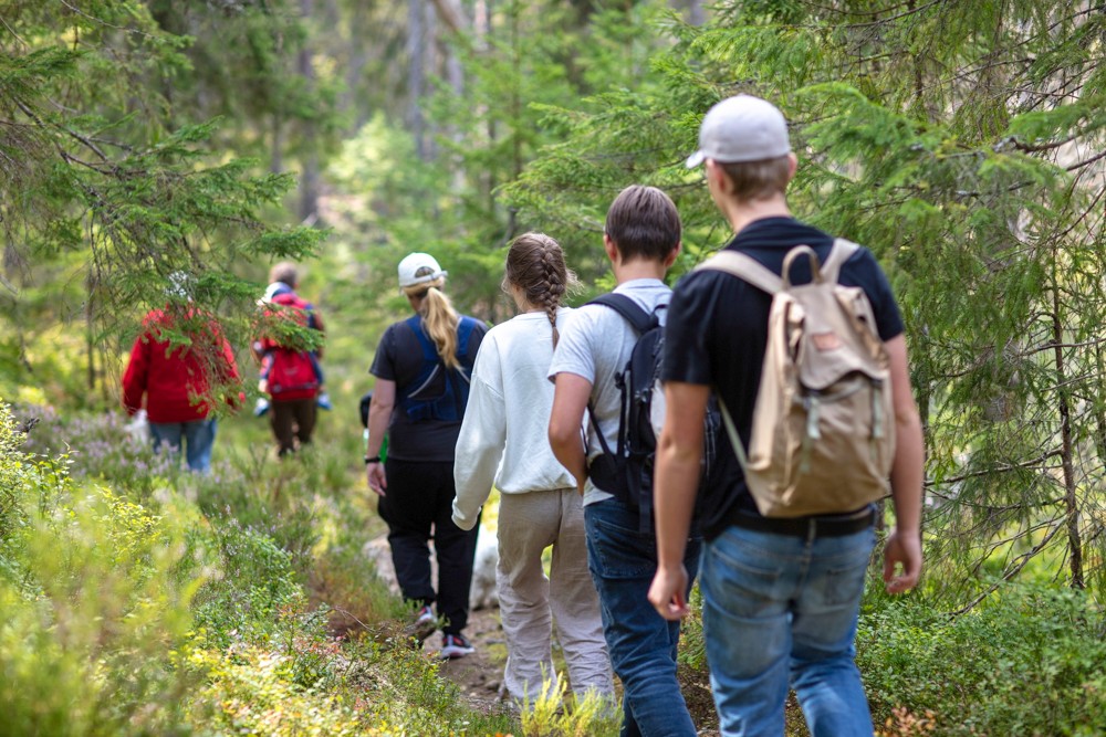 Människor i olika åldrar går på rad genom skogen. Flera bär ryggsäck. 