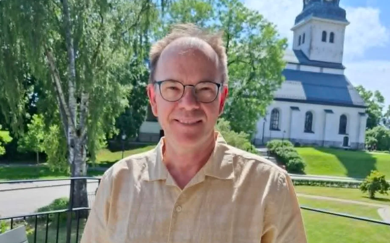 En medelålders man i halvfigur står framför en vit kyrka.