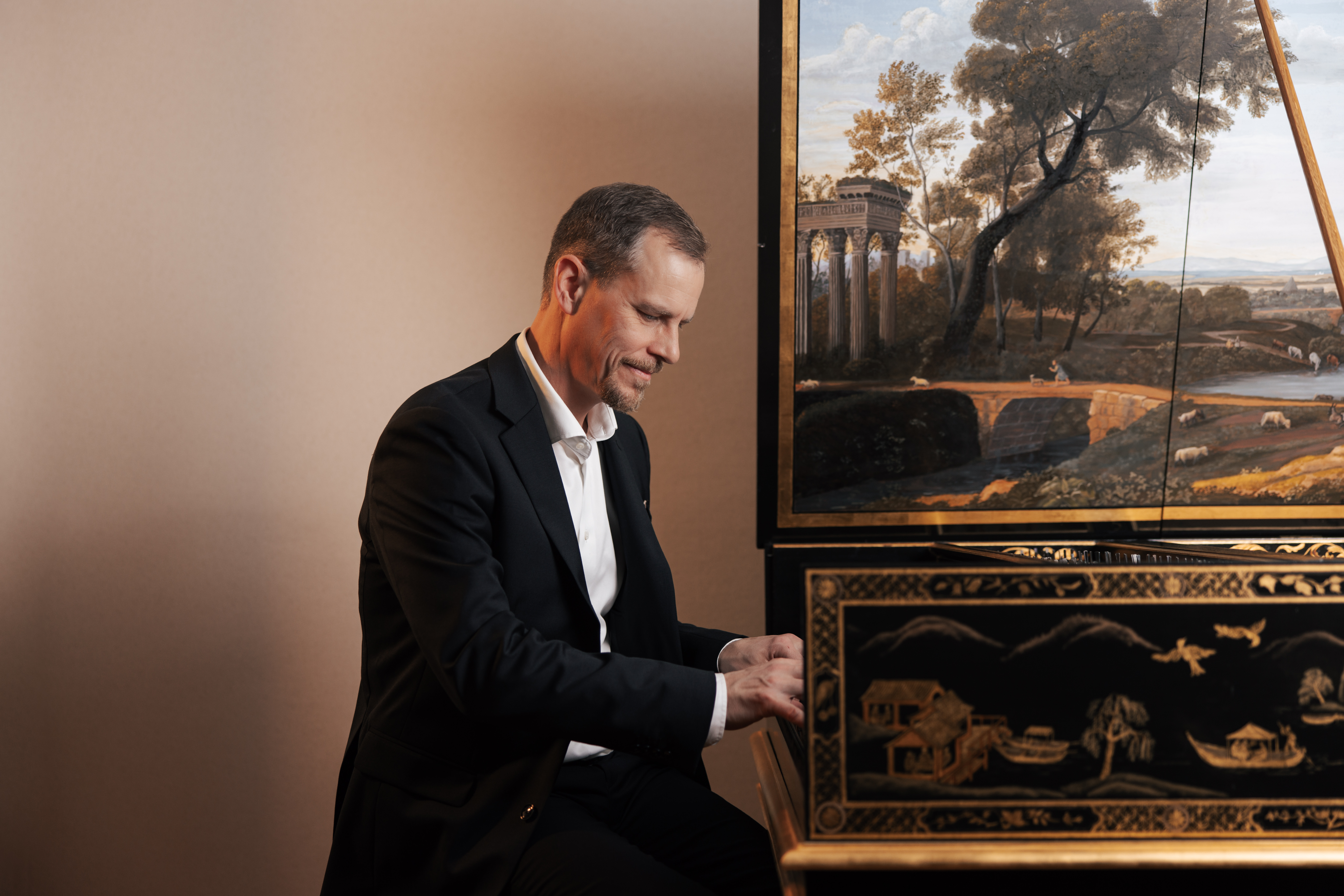 En man med svart kostym sitter vid ett piano, I bakgrunden finns en stor oljemålning på ett träd och vita, smyckade pelare.