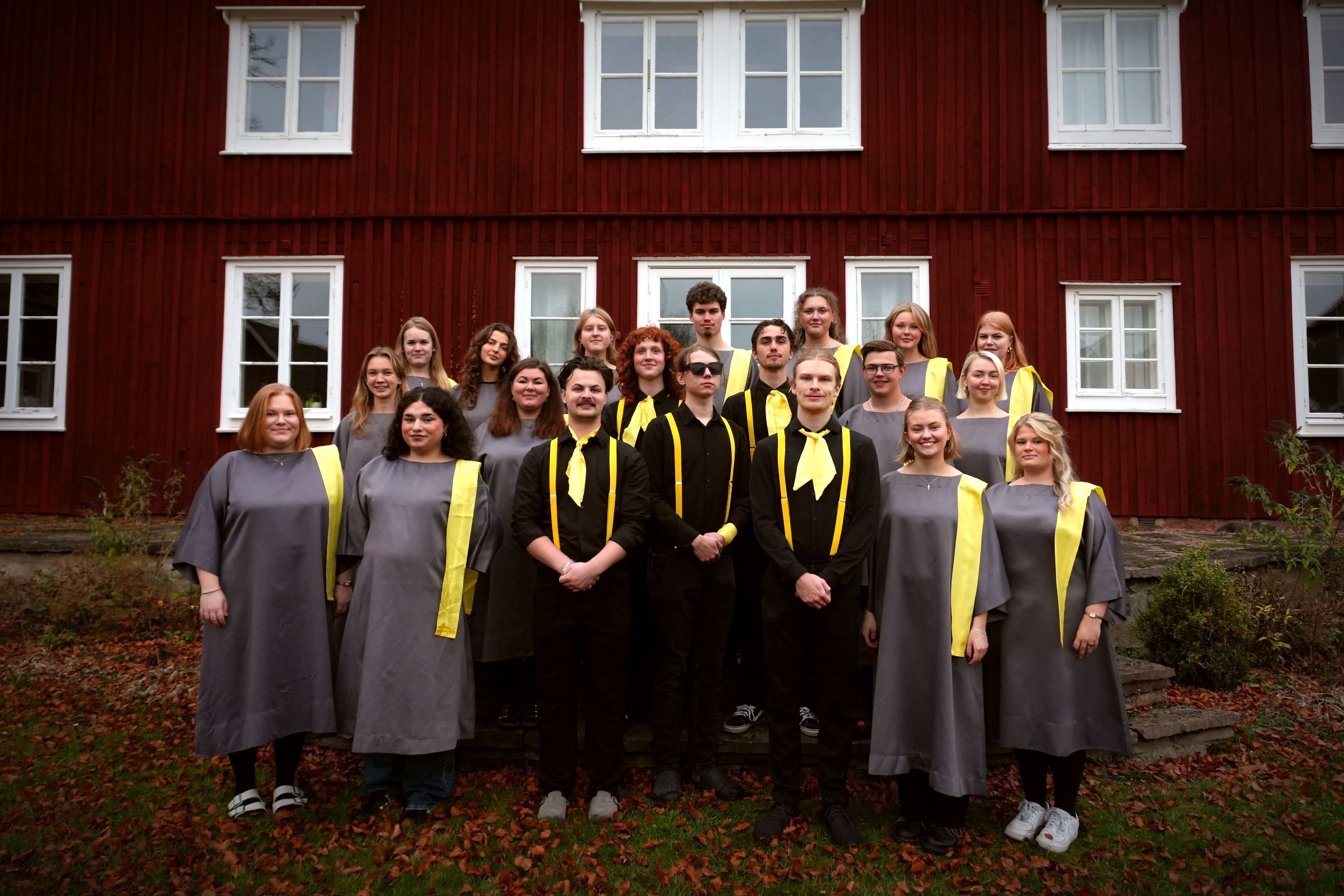 En grupp människor med gråa och svarta gospelkläder med gula detaljer står framför ett falurött hus med vita knutar.