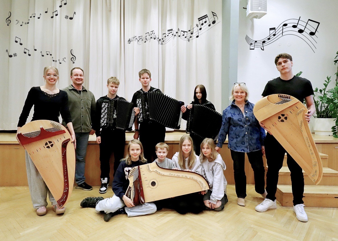 Ryhmä eri-ikäisiä nuoria ja aikuisia seisoo ja istuu lavalla musiikkisalin edessä. He pitävät käsissään erilaisia kansansoittimia, kuten kanteleita ja harmonikkoja. Taustalla on vaalea verho, jota koristavat nuottikuvat. Ryhmä poseeraa yhdessä rennossa tunnelmassa.