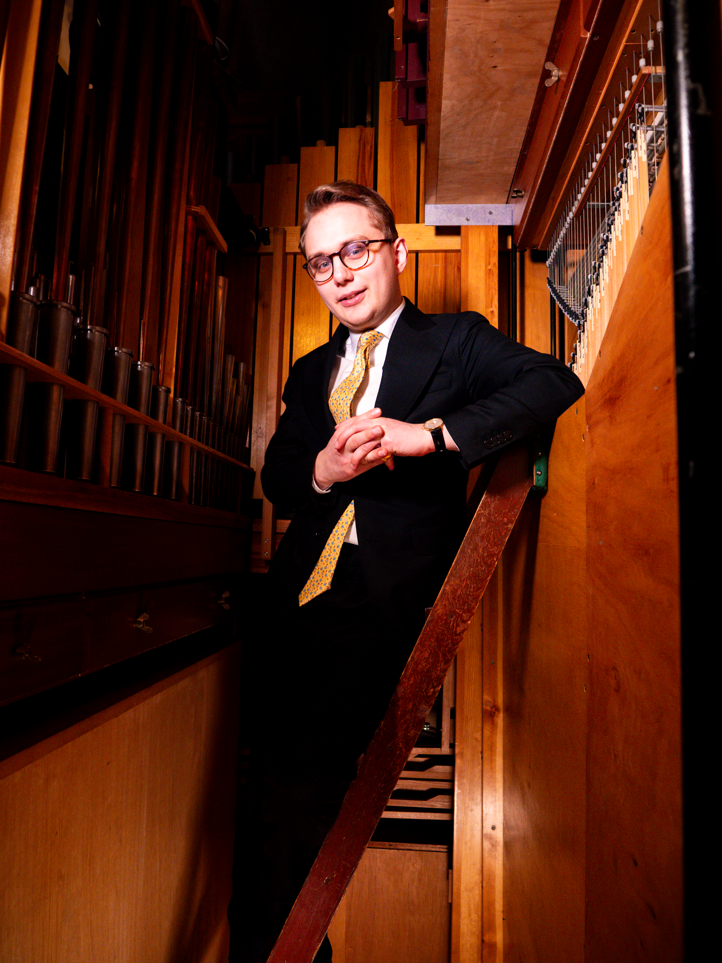 En ung man står lutad mot en trävägg i en trång orgel. Han har kostym och gul slips.