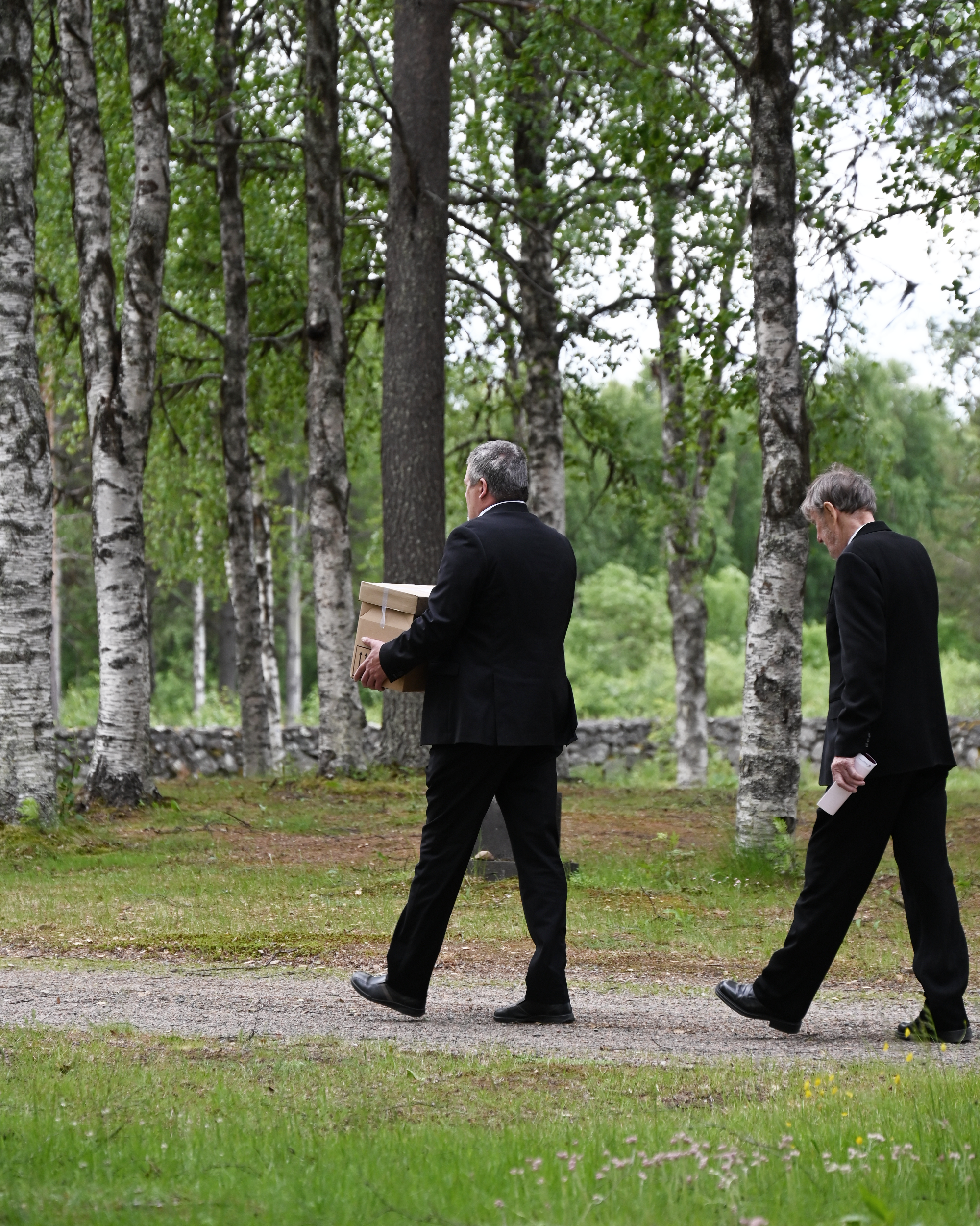 Två män i svarta kläder går på en kyrkogård. En av dem bär en pappkartong.