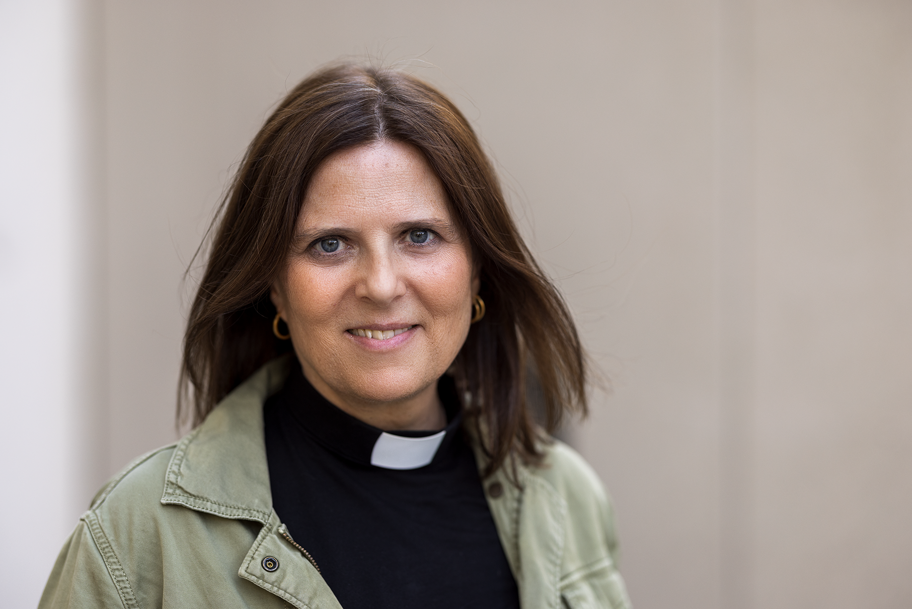 Leende kvinna, brunt hår, prästkrage runt halsen mot grå bakgrund 