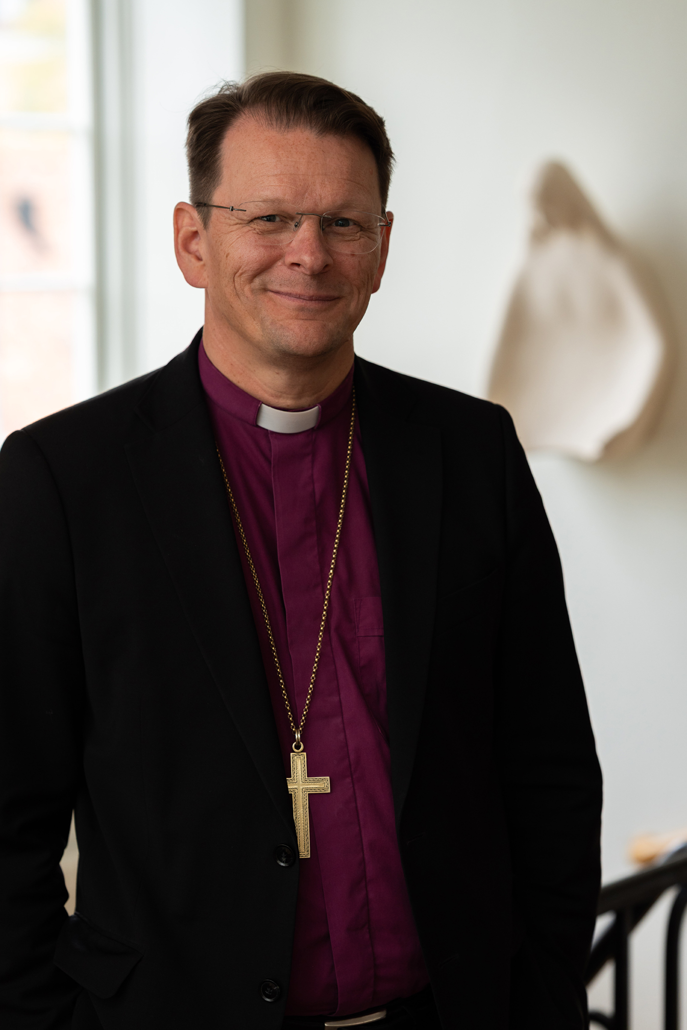 Biskop Erik står i trappan framför ett fönster i Biskopgården klädd i lila biskopsskjorta och biskopskorset kring halsen. 