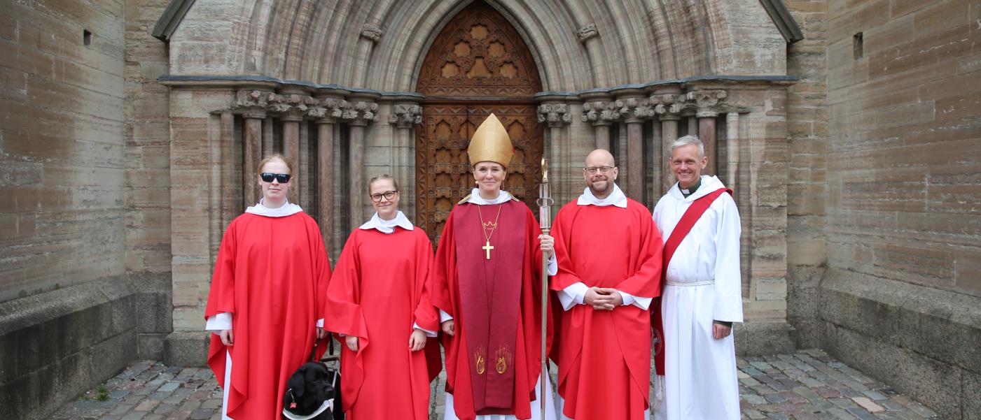 Tre nyvigda präster, en nyvigd diakon, en ledarhund och en biskop står utomhus framför en kyrkport.