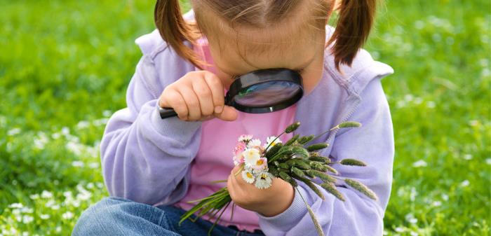 En liten flicka sitter i gräset och ser på blommor genom ett förstoringsglas.