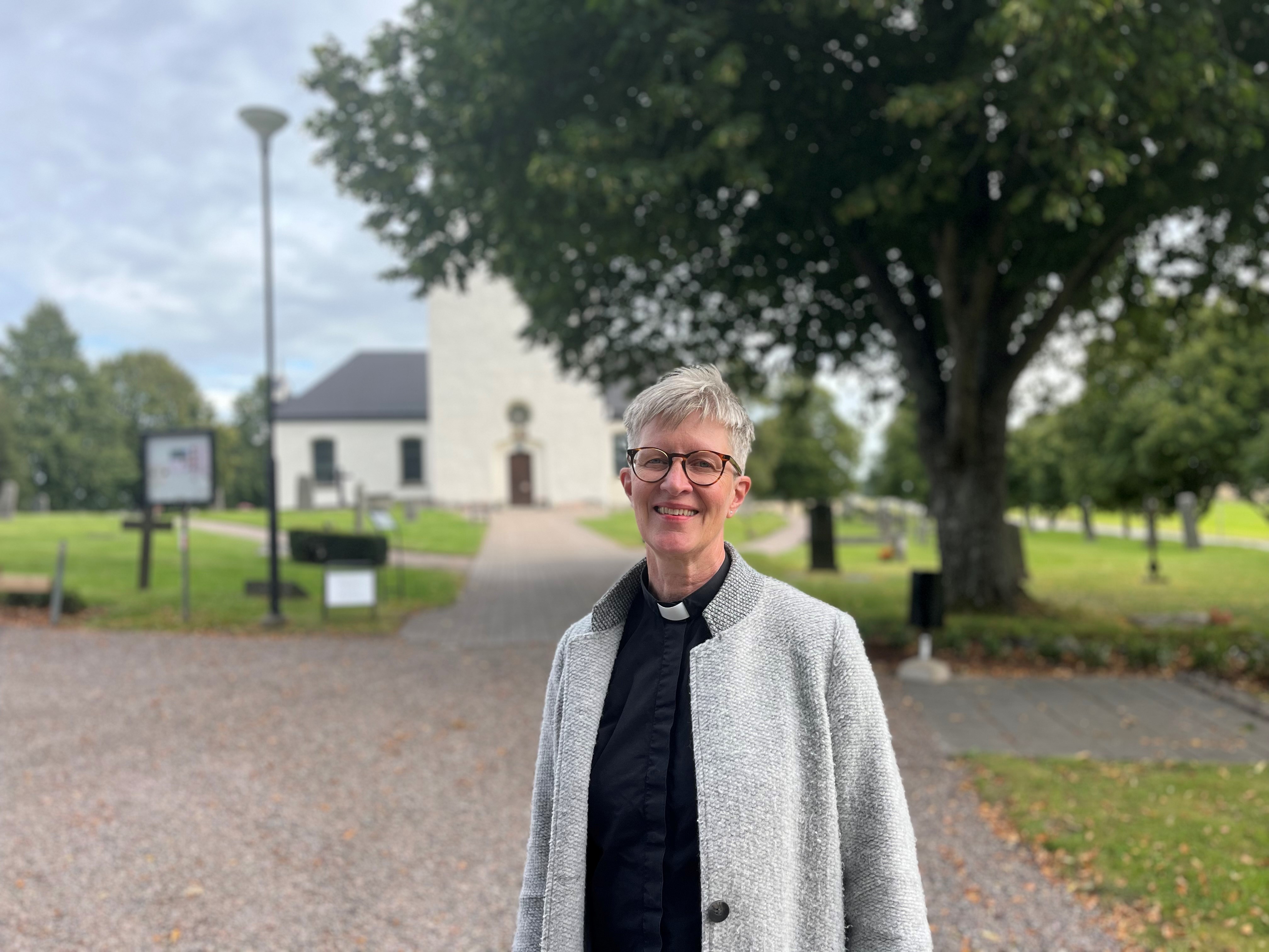 Kvinna i prästkläder som står framför vitkalkad kyrka och kyrkogård.