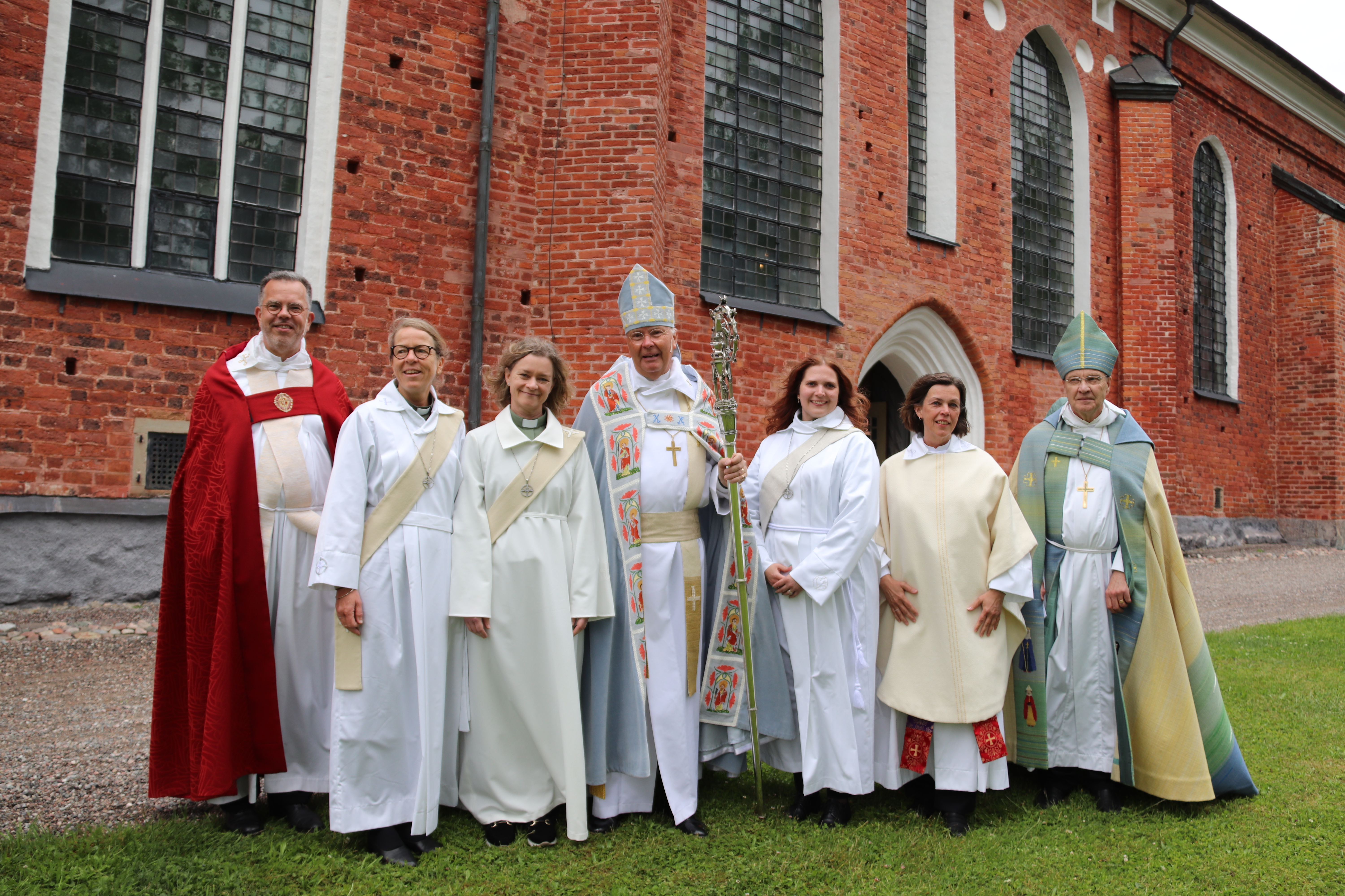Sju personer: präster, diakoner och biskopar i liturgiska kläder framför katedral i rött tegel.